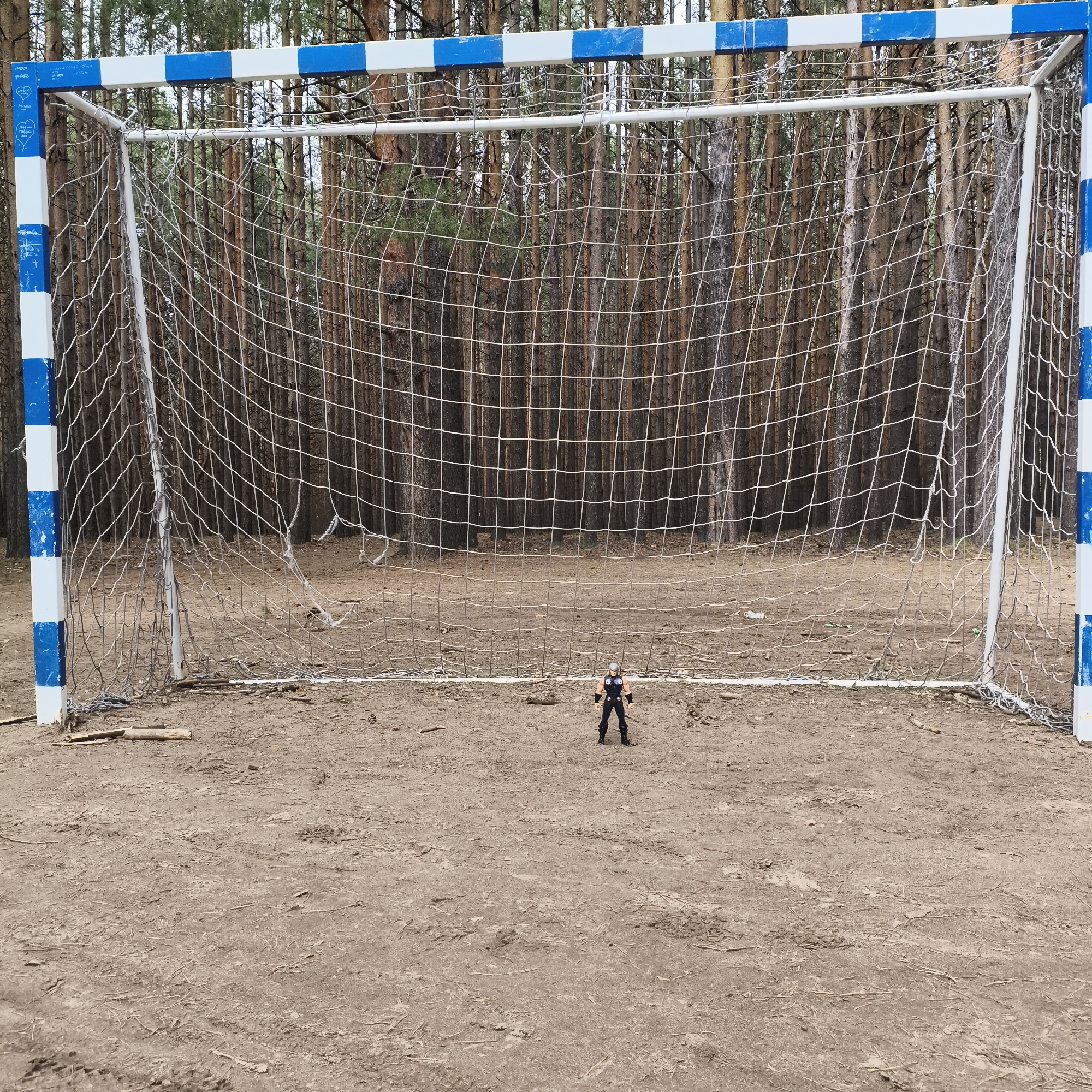 The image depicts a soccer goal situated on a dirt field in front of a pine forest. The goalposts are painted with alternating blue and white stripes. A white net is stretched across the goal frame. In the center of the field, in front of the goal, stands a small Thor action figure. The scene appears to be located near a forest in Chaikovsky, Russia.