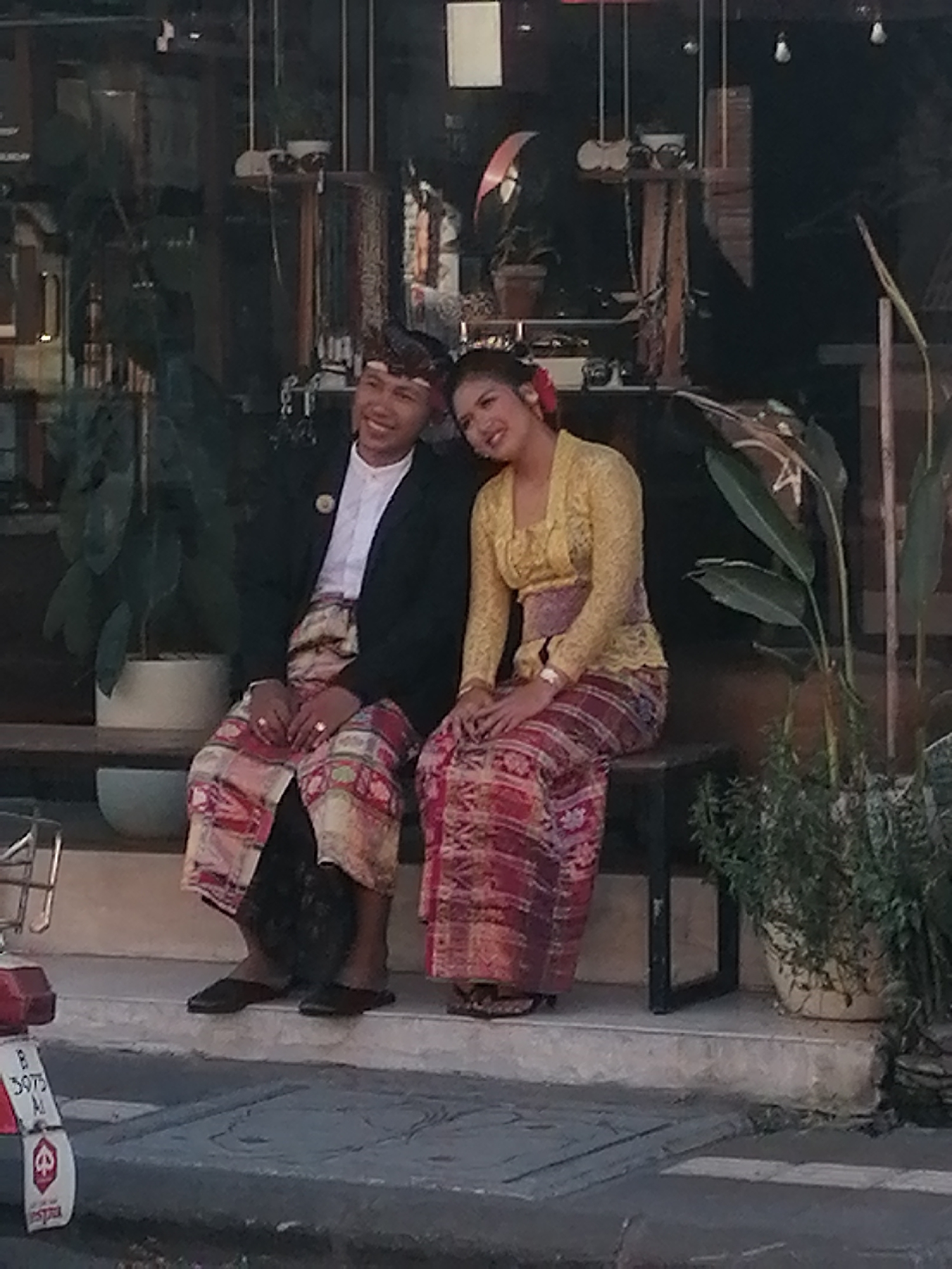An adult male and an adult female, dressed in traditional Balinese attire, are seated on a bench on an elevated platform. The male wears a black jacket over a white shirt and a patterned sarong. He has a traditional headpiece and rings on his fingers. The female wears a yellow lace top with a patterned sash and a patterned sarong. She has a red flower in her hair. They are posed together, with the female leaning her head on the male's shoulder, and both are smiling. Behind them, a storefront is visible with displays of merchandise and plants. In the foreground, a portion of a red and white moped is visible, with a license plate that reads "B 3975 AJ". The scene appears to be outdoors in a public area, possibly a shop or restaurant in Ubud District, Indonesia.