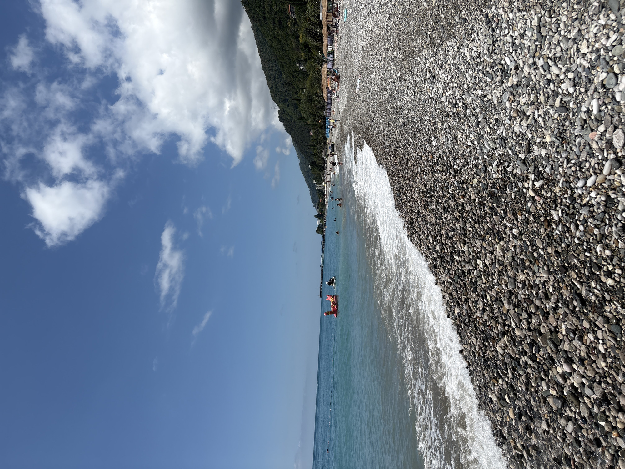 A wide-angle, eye-level shot captures a scenic view of a pebble beach and the Black Sea under a partly cloudy sky. The foreground is dominated by the textured surface of the beach, composed of smooth, grey, and dark-colored pebbles. Gentle waves roll onto the shore, creating a foamy white border along the water's edge. The sea itself is a vibrant turquoise, appearing calm with some small ripples.
In the water, a few people are visible. An inflatable red and yellow float, shaped like an animal, is present. A jet ski is also in the water, with a person riding it. Further out, a long pier extends into the sea, with several people on it.
The background reveals a lush green mountainside densely covered with trees, suggesting a tropical or subtropical climate. The slopes are dotted with buildings and some visible utility poles. Along the shoreline, a row of beach umbrellas with thatched roofs provides shade for beachgoers. Several individuals are seen relaxing on lounge chairs or walking along the beach. The scene conveys a sense of a popular tourist destination, likely during the daytime in warm weather.