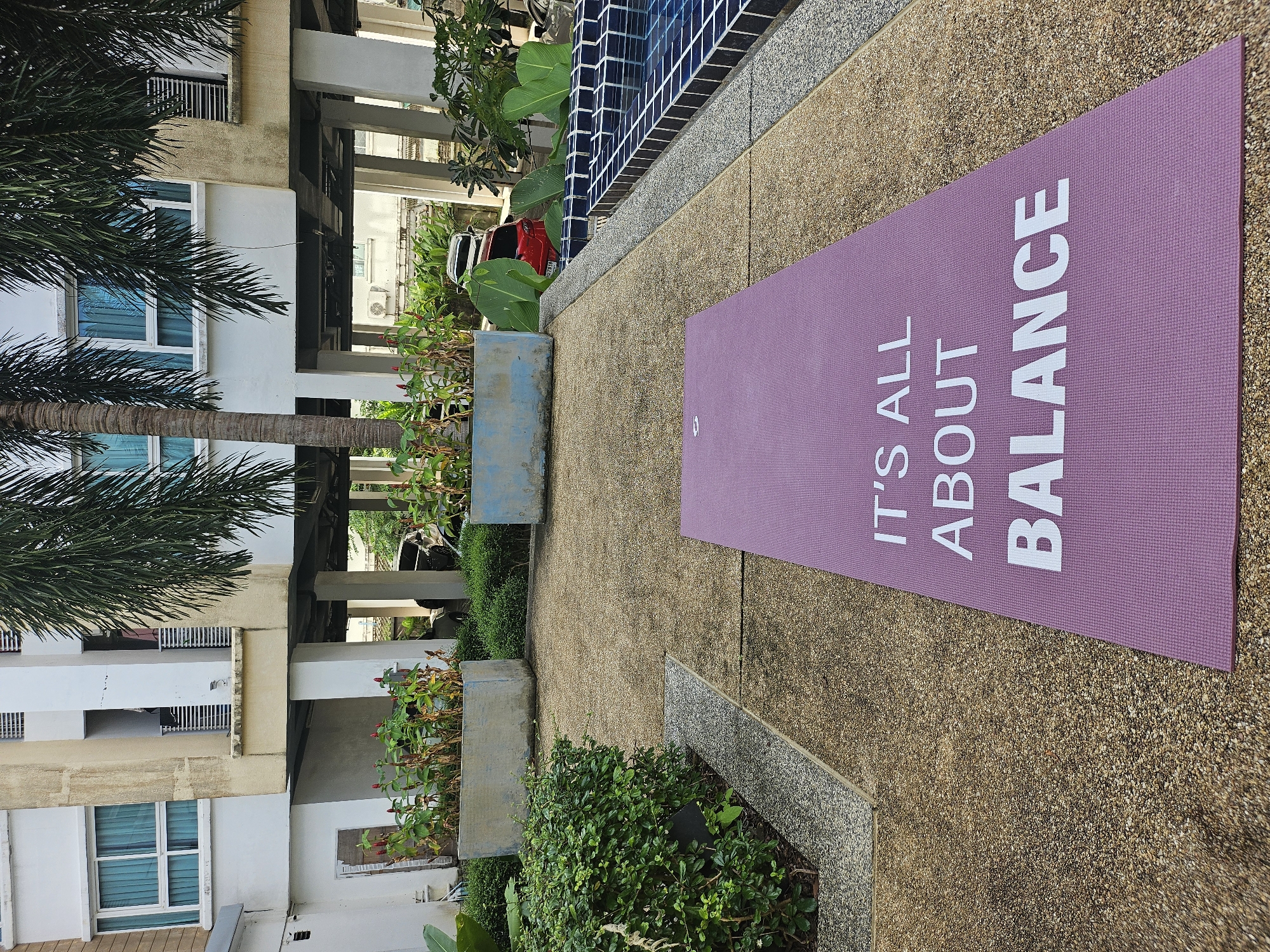 A purple yoga mat is laid out on a pebble-textured walkway in front of a residential building in Kathu, Thailand. The building has multiple stories with cream-colored walls and white pillars. Several palm trees with lush green fronds are visible, casting shadows on the walkway. Green bushes and planter boxes with red-flowered plants are arranged around the area. To the right of the mat, a portion of a tiled swimming pool is visible. The mat has white text printed on it, reading "IT'S ALL ABOUT BALANCE". In the background, parked cars can be seen beneath the building's elevated structure.