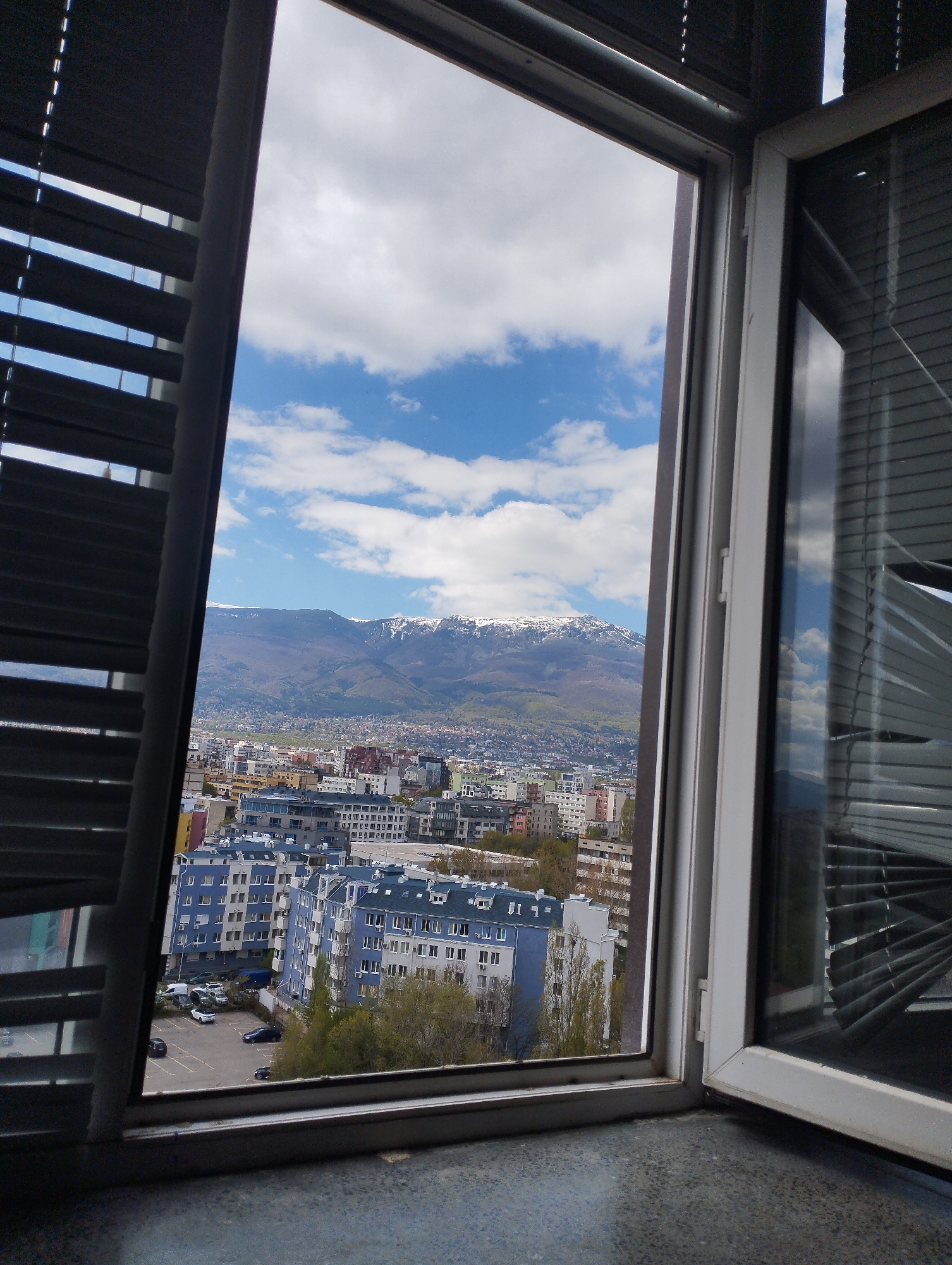 The image presents a vibrant daytime view of a city against a backdrop of majestic, snow-capped mountains, all framed by an open window.
In the immediate foreground, the dark grey frame of an open window occupies the center, with a textured dark window sill extending across the bottom. On the left side, dark Venetian blinds are partially drawn, allowing glimpses of the sky through their slats. Similarly, on the right side, another pane of the window shows reflections of the sky and adjacent buildings, with similar dark blinds partially visible.
Beyond the window, a bustling urban landscape stretches out. Numerous multi-story buildings, predominantly residential or commercial, fill the midground. Several buildings are notable for their distinct blue and white facades, while others are in various shades of grey, white, and even hints of pink. A parking lot with several parked cars (including white, dark blue, and black vehicles) is visible among the buildings, surrounded by trees with light green foliage, suggesting spring or early summer.
In the distance, a range of mountains rises prominently. Their peaks are dotted with patches of white snow, indicating either high altitude or a cooler climate, contrasting with the green and brown tones of their lower slopes.
The sky above is a clear, brilliant blue, adorned with scattered, fluffy white clouds. The bright, natural lighting indicates that the photograph was taken on a sunny day, likely during late morning or early afternoon. No specific human activity is discernible, and no visible text is present on any buildings or signs. The scene evokes a feeling of calm observation of a lively city coexisting with grand natural scenery. The specific geographical location is not identifiable from the image.