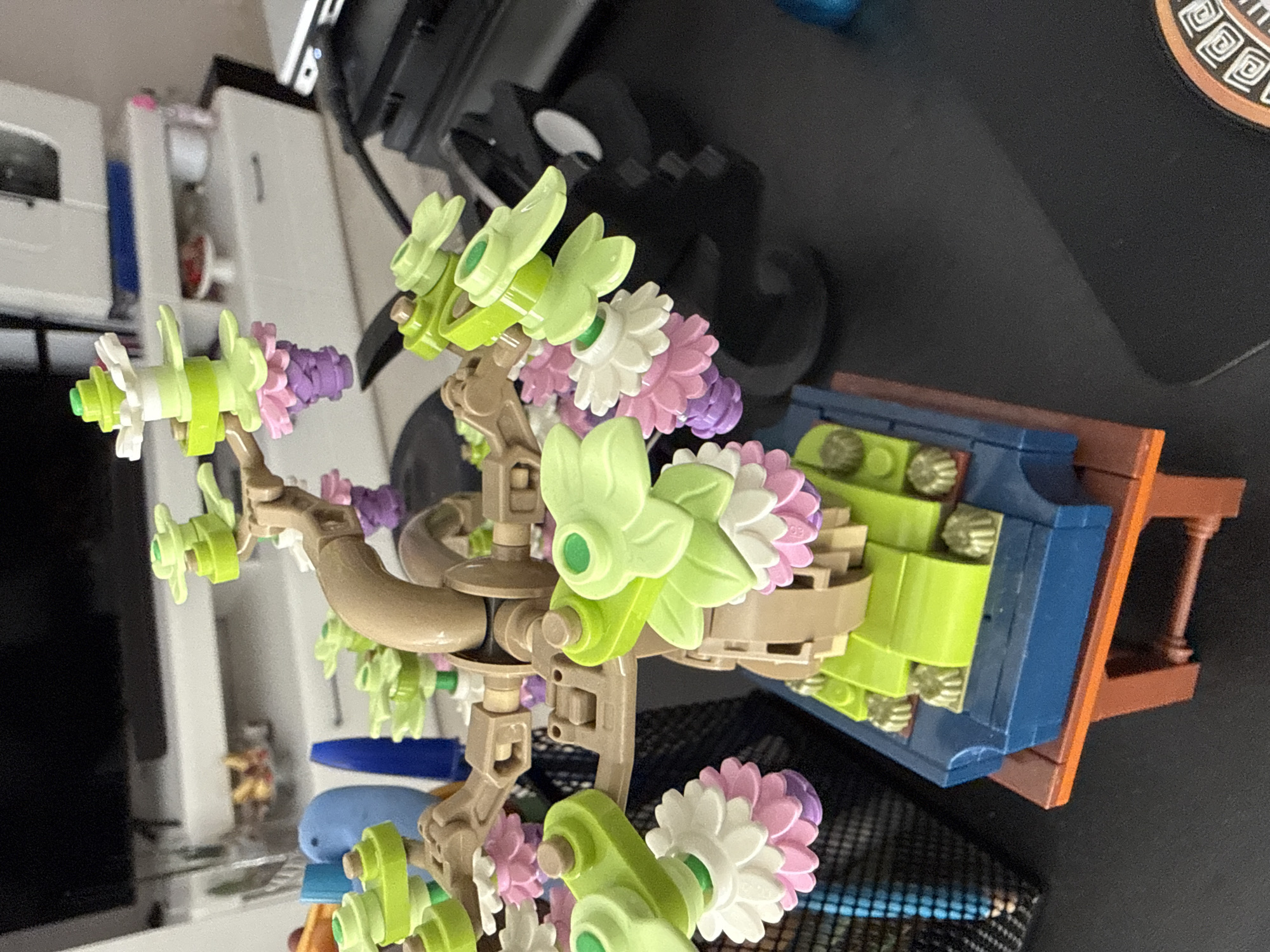 The image shows a close-up of a LEGO flower tree model placed on a dark surface, possibly a desk. The tree has light brown branches and is adorned with various colorful LEGO pieces representing leaves and flowers in shades of green, white, pink, and purple. The base of the tree is a blue LEGO planter with green elements resembling grass or foliage, all resting on an orange LEGO stand. In the background, out of focus, there is a white cabinet with drawers and a black electronic device, suggesting an indoor setting. There are no people or animals visible in the image. The image does not provide any information about the city of Dobrich, Bulgaria.