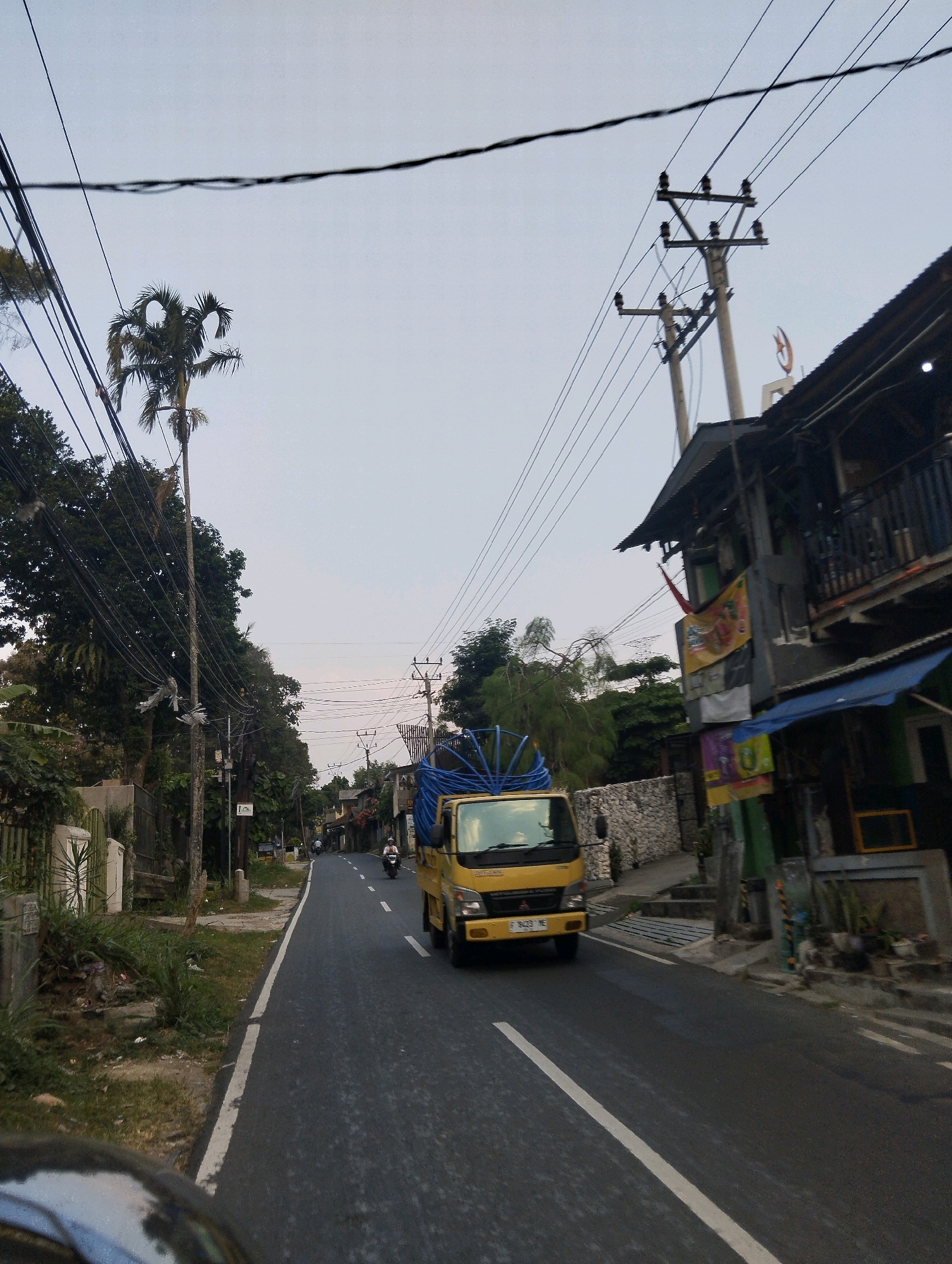 The image depicts a street scene in Megamendung, Indonesia. A yellow truck with a blue tarpaulin covering its cargo is traveling down a paved road. The truck's license plate reads "F 9423 ME". On the right side of the road, a multi-story building with a dark-colored roof and balconies is visible, adorned with several banners and signs. A power pole with multiple electrical wires runs along the street. On the left side, there are trees, a fence, and more electrical poles and wires. A motorcyclist is also seen on the road, traveling in the same direction as the truck. The sky is overcast with a pale gray hue, suggesting it may be early morning or late afternoon. The environment appears to be a semi-urban or residential area.