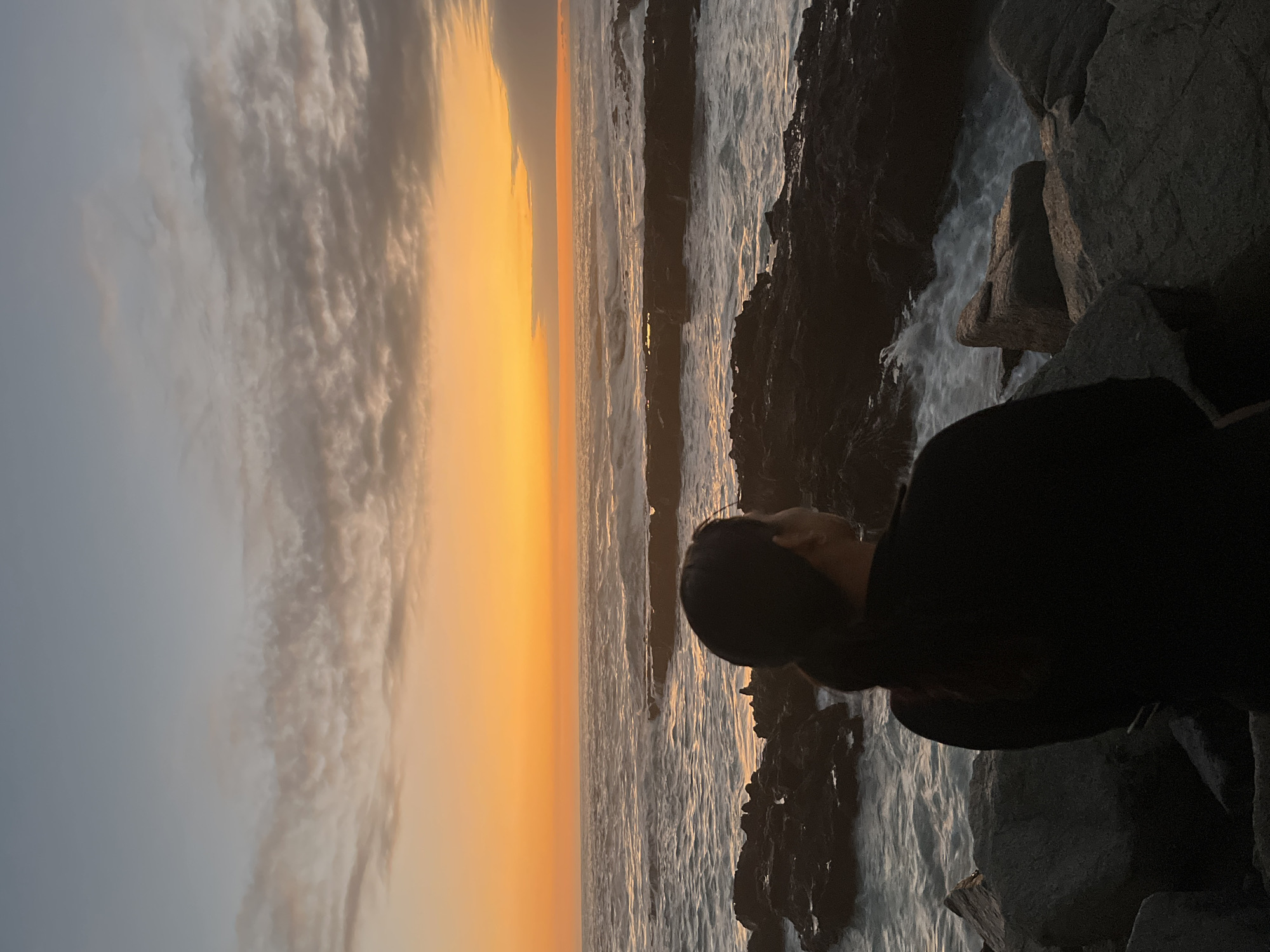 The image, captured in a vertical orientation, depicts a serene coastal scene at either sunrise or sunset. A person, viewed from behind and slightly to the right, is the main human subject, their dark silhouette facing the expansive ocean. They appear to be sitting or standing on a rocky outcrop along the shore.
The sky dominates the upper portion of the frame, transitioning from a soft grey-blue at the top to a vibrant, warm orange and yellow along the horizon, indicative of either dawn or dusk. Wispy, stratified clouds stretch across the sky, catching the light and adding texture.
The ocean occupies the middle ground, its surface reflecting the sky's colors, appearing a mix of deep blues, greys, and shimmering oranges where the light hits the water. Waves are visible, with white foam and turbulent water near the rocky shore, suggesting the force of the sea.
In the foreground and to the right, dark, rugged rocks form the coastline, creating a natural barrier between the observer and the open water. These rocks are shadowed, enhancing the contrast with the bright horizon and highlighting the person's silhouette. The overall mood is one of contemplation and appreciation for nature's beauty during a transitional time of day. No visible text is present in the image.