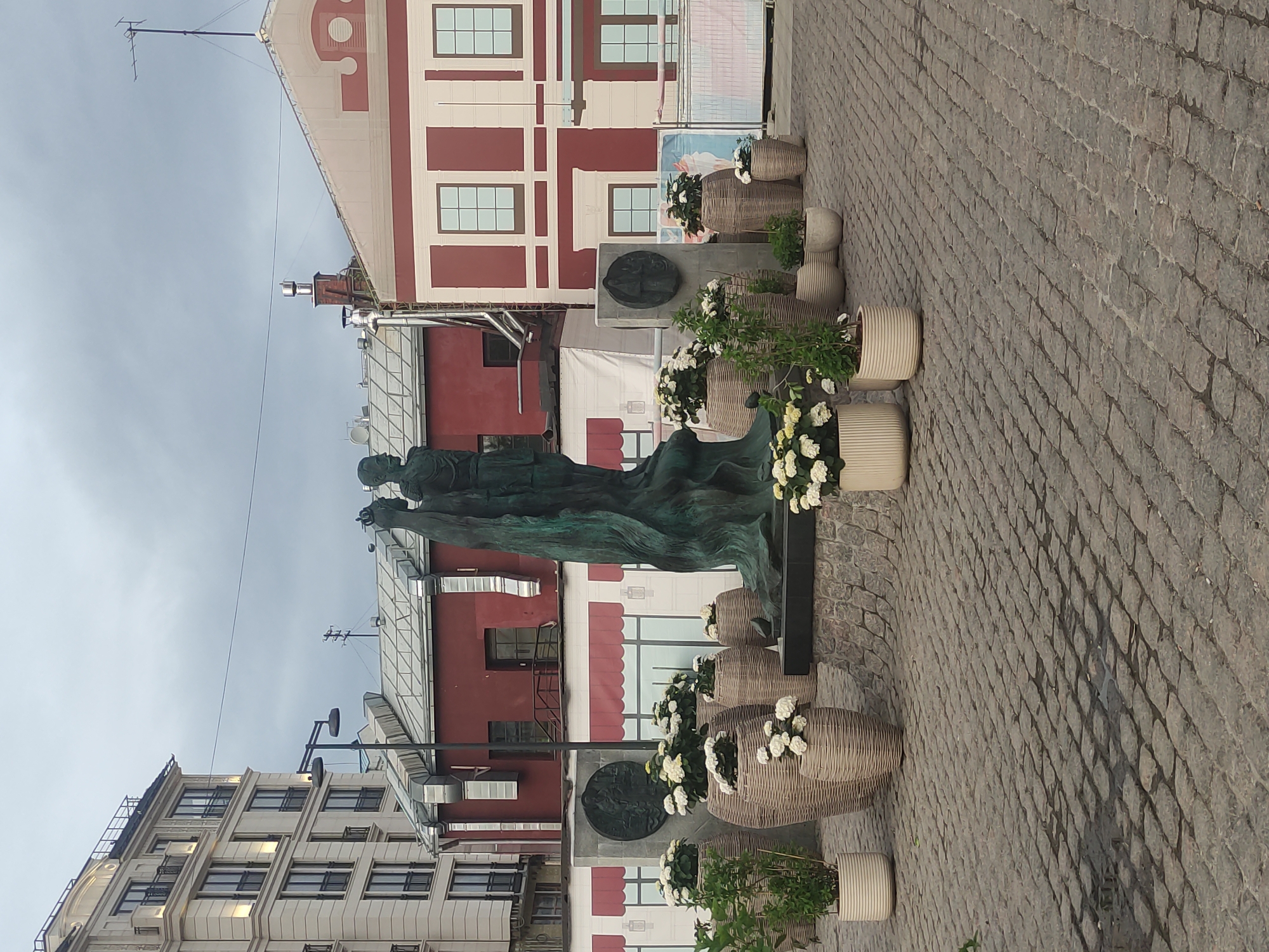 The image shows a statue in Moscow, Russia. The statue depicts two figures and is surrounded by flowerpots and brickwork. In the background, there are buildings of varying heights and styles. The sky is overcast. The ground is paved with cobblestones.