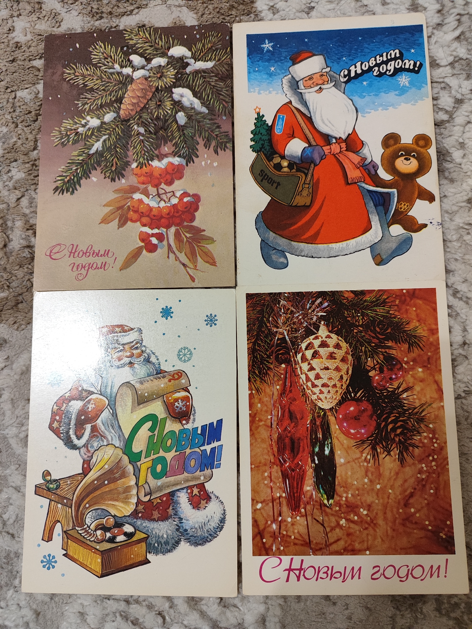 The image displays four vintage Soviet-era New Year's postcards arranged in a 2x2 grid. The postcards are placed on a textured grey surface that appears to be a carpet or blanket.
The top-left postcard features a snow-covered spruce branch with a pine cone and bright red rowan berries. The text "С Новым годом!" (Happy New Year!) is written in a cursive pink font on a tan background at the bottom left.
The top-right postcard depicts a smiling Father Frost (Ded Moroz) in a red coat, a blue hat, and a white beard, holding a bag labeled "Sport" filled with a soccer ball and other items. Next to him is a brown bear, possibly representing the Olympic mascot Misha. The text "С Новым годом!" is written in a stylized font in the upper right corner against a blue, starry sky.
The bottom-left postcard shows Father Frost, again in traditional attire, holding a scroll with the text "С НОВЫМ ГОДОМ!" (Happy New Year!) in large, colorful letters. In front of him is a wooden gramophone and a small wooden stool with a jewelry box. Blue snowflakes are scattered around the scene.
The bottom-right postcard is a close-up of a decorated fir tree branch with various ornaments, including a large, textured pine cone ornament, elongated red and green glass ornaments, and red apple ornaments. The background is a warm, bokeh-like blur of lights. The text "С Новым годом!" is written in a cursive pink font across the bottom.
The image does not contain any elements that would indicate its location in Pavlodar, Kazakhstan. The scene is a collection of holiday-themed postcards.