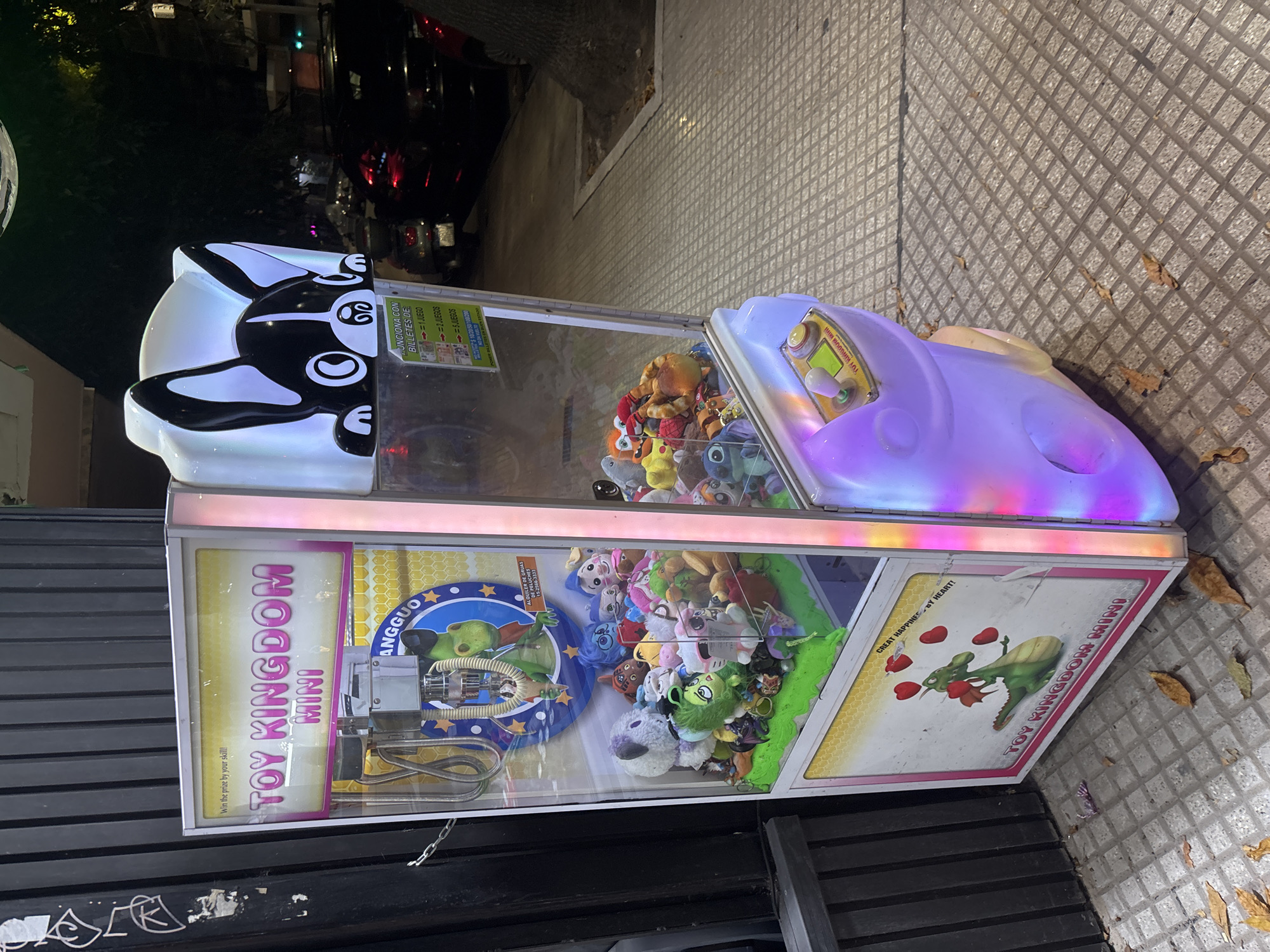 The image shows a "Toy Kingdom Mini" claw machine on a sidewalk in what appears to be a commercial area. The machine features a black and white cartoon dog design on the top and is filled with plush toys. Signage on the machine advertises the games and mentions "Alquiler de gruas de peluches 15-2566-3333" and says it's "Funciona con billetes de". The sidewalk is tiled with brown autumn leaves scattered around. In the background, there is a street scene with cars and trees, indicating an urban setting. Since the phone number is from Buenos Aires, and the language on the machine is Spanish, the location is very likely in the city of Buenos Aires, Argentina.