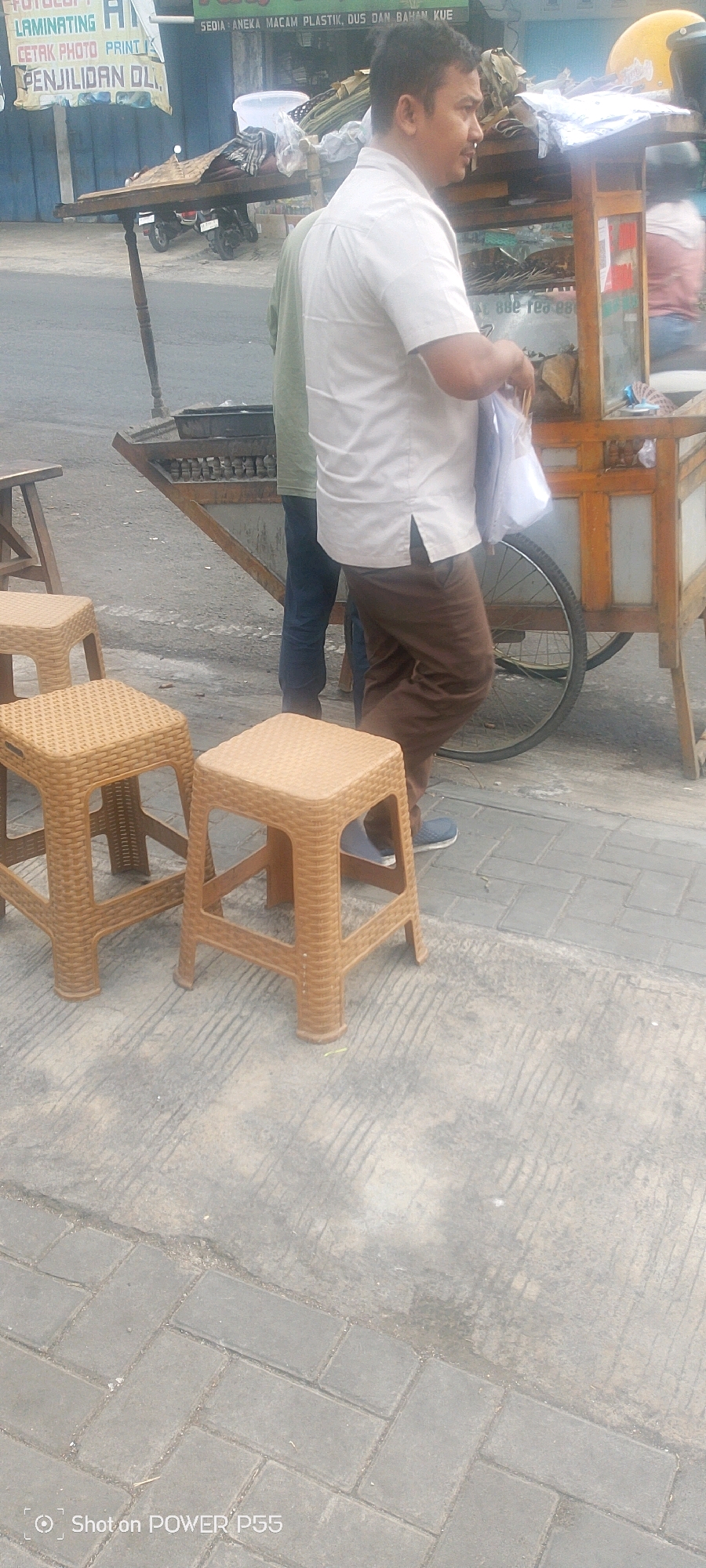 An eye-level, medium shot captures a street scene in an urban environment. In the foreground, three tan, woven plastic stools are arranged on a paved walkway. Behind the stools, a man in a light-colored short-sleeved shirt and brown pants stands next to a food cart. He is holding a white plastic bag and appears to be interacting with someone off-camera or with the cart. Another person, wearing a green shirt and blue jeans, stands to the left of the man, partially obscured by him. The food cart is made of wood and metal, with a wheel visible on the right side. In the background, a street with parked motorcycles is visible, along with banners advertising various services like "LAMINATING," "CETAK PHOTO PRINT," and "PENJILIDAN OL." The overall environment suggests a commercial area. The lower left corner of the image contains a watermark indicating "Shot on POWER P55."
