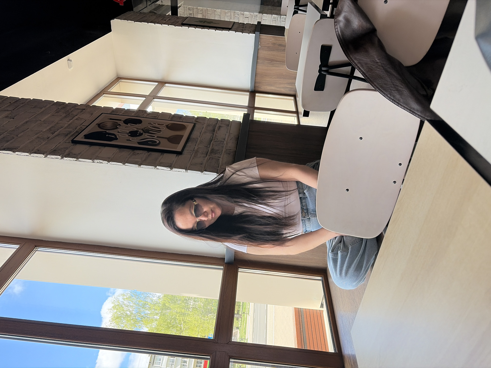 A woman with long dark hair and sunglasses is sitting at a table in a restaurant. She is wearing a white t-shirt and blue jeans. The table is light brown and there are white chairs around it. There is a large window behind her with a view of a blue sky, green trees, and buildings. A black and white picture hangs on the brick wall to the left of the woman. A brown leather jacket is draped over the back of a chair to the right of the woman. The location appears to be an urban area, in the city of Unknown, Unknown.