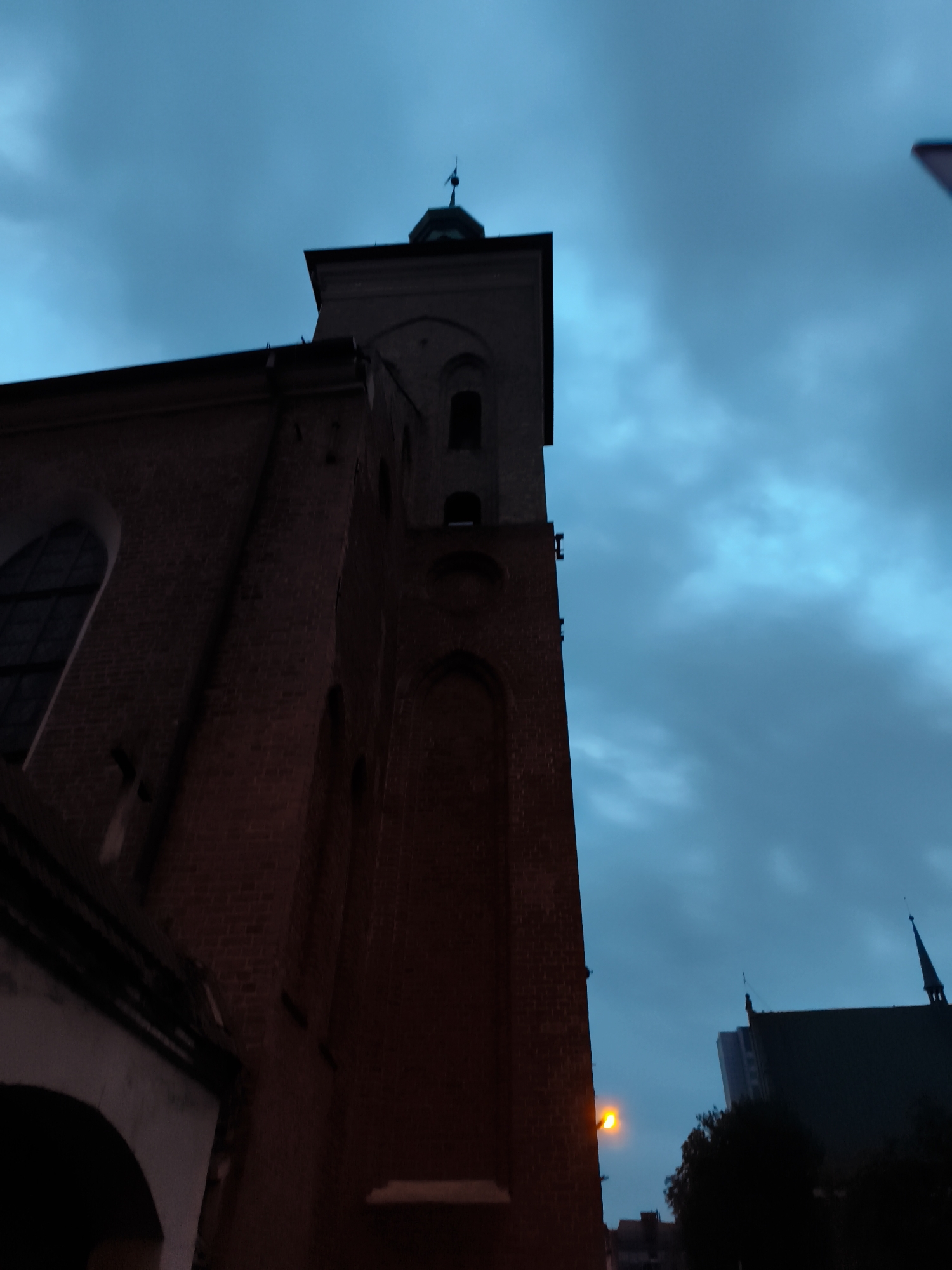 The image shows a low-angle view of a brick church tower against a dark blue, cloudy sky. The tower is part of a larger brick building on the left of the frame, with a large arched window visible. In the background on the right, a dark roofline with spires is visible, along with a tall modern building behind it. A street lamp with a yellow light is also visible on the left, near the church building. The overall impression is of a dimly lit urban scene, possibly at dusk or dawn. The church is St. Mary's Church in Gdańsk, Poland.