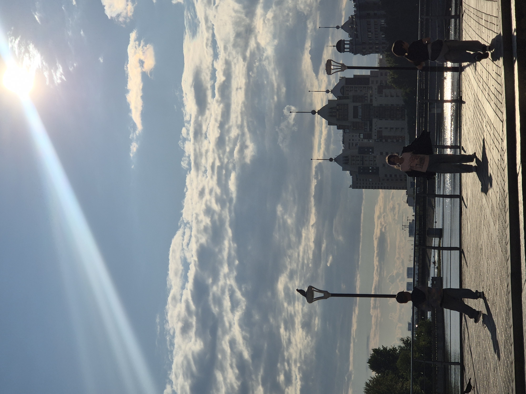 This is a photograph of the waterfront area of Astana, Kazakhstan, likely taken in the late afternoon or early evening given the strong backlight from the sun. In the foreground, three children are silhouetted against the bright sky and water. Two children appear to be walking or running on a paved walkway, with the third child further to the left, facing away from the camera. A bird is perched on a street lamp on the left side of the image, and another bird is on the ground in the lower left corner. In the mid-ground, a body of water, possibly the Ishim River, is visible, with a railing separating the walkway from the water. Beyond the water, several modern buildings with distinctive architectural features, including domed roofs and spires, are visible, characteristic of Astana's cityscape. The sky is filled with dramatic clouds, illuminated by the setting sun, which casts a bright ray across the upper portion of the image. The overall atmosphere is one of leisure and urban scenery under a striking sky.