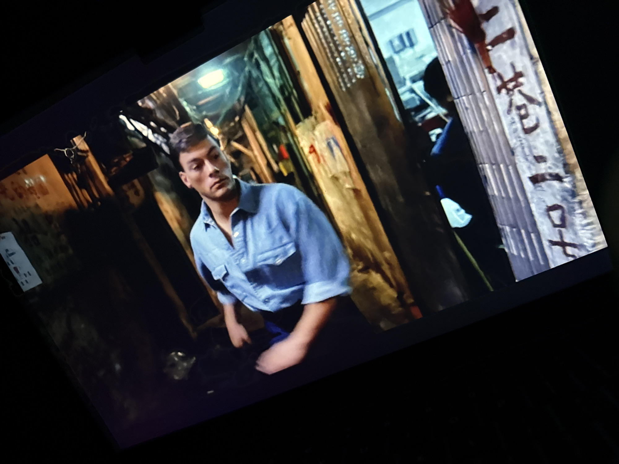 The image shows a scene from what appears to be a movie or TV show. A man with short brown hair, wearing a blue short-sleeved button-down shirt and dark pants, stands in a narrow, dark alleyway. The alley has wooden supports or walls with writing in what looks like Chinese script on some of them. The environment appears gritty, potentially an urban backstreet location, possibly in an Asian setting based on the script and overall atmosphere. There is an open area beyond the alley where a blurred figure in a dark uniform is visible. The man seems to be observing or moving towards the scene unfolding in the background. This scene is not in Plovdiv, Bulgaria.