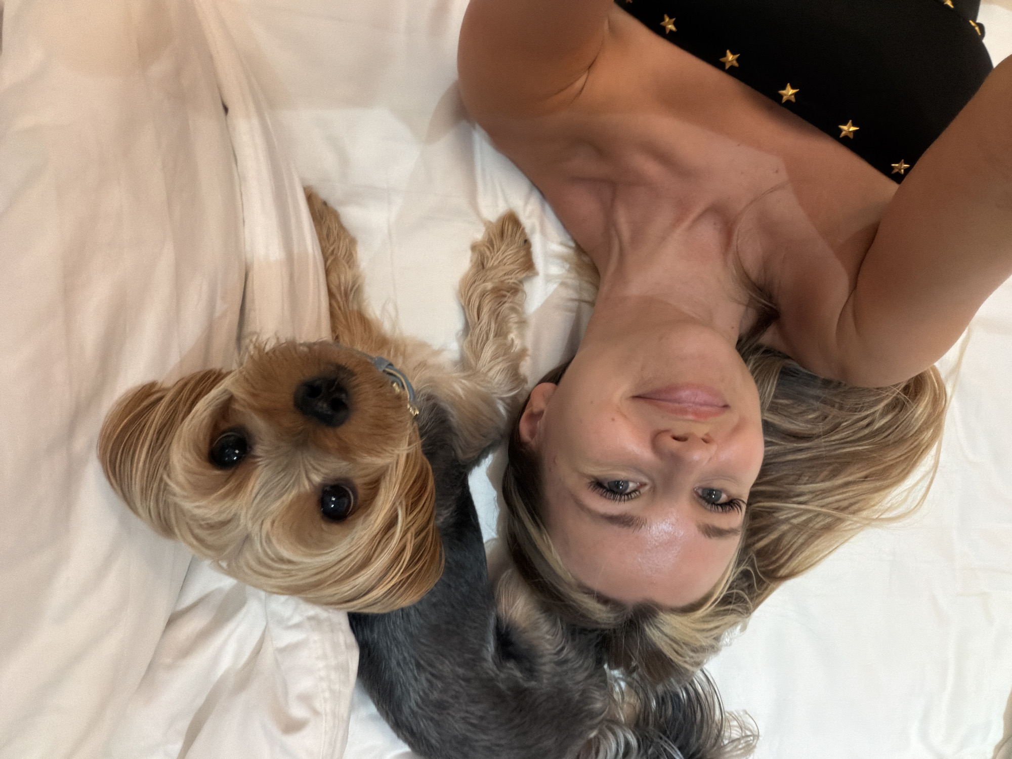 A woman and a Yorkshire Terrier are lying on a white, rumpled bed, possibly in a hotel room in Thep Krasatti, Thailand. The woman is lying on her back, facing the camera and smiling. She has blonde hair that cascades around her face and shoulders. She is wearing a black top with gold stars on it. The Yorkshire Terrier is positioned to the left of the woman, looking directly at the camera with large, dark eyes. Its fur is a mix of tan and grey. The scene appears to be indoors, and the lighting suggests it could be daytime. The mood is relaxed and affectionate, with both subjects looking comfortable and content. There is no visible text in the image.
