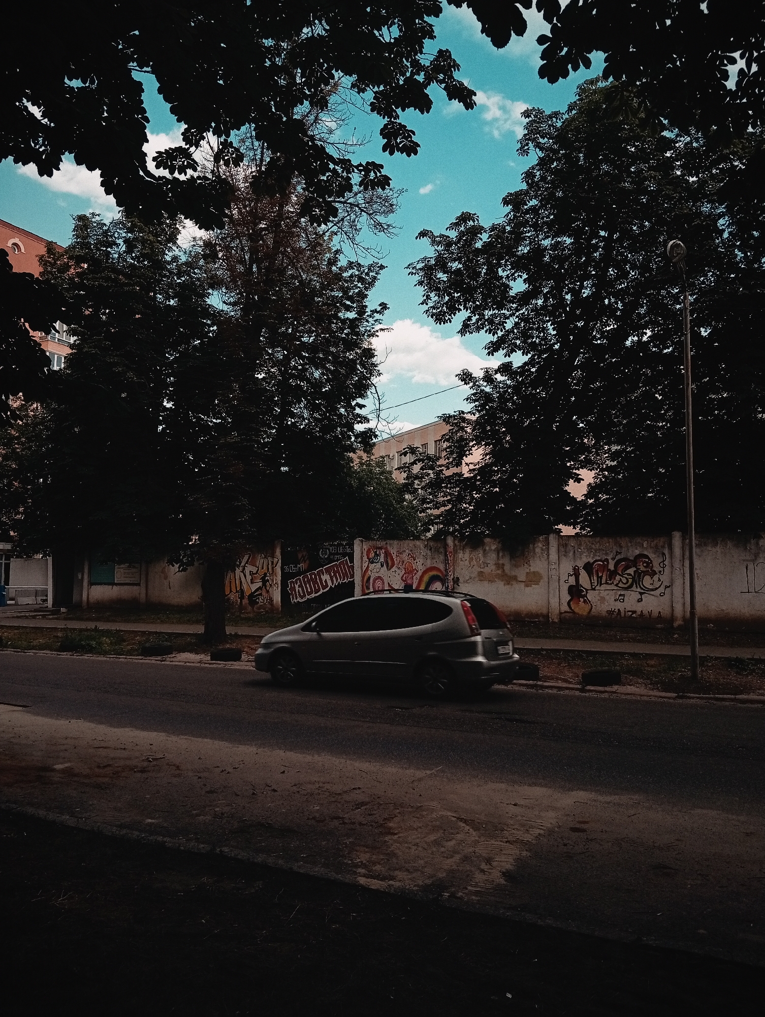 The image depicts a street scene with a car driving past a concrete wall covered in graffiti. The car is a silver hatchback, viewed from the side, moving from left to right. The wall is weathered and adorned with various colorful spray-painted designs, including what appears to be a rainbow and abstract lettering.
Tall, dark trees with dense foliage frame the top and sides of the image, casting shadows and creating a somewhat dramatic contrast with the bright blue sky dotted with white clouds visible between the branches. Behind the wall, parts of buildings are visible, including a peach-colored building on the left and a lighter colored building further back. A street lamp stands on the right side of the image. The road in the foreground is asphalt, with some debris and patches of lighter-colored material.
The lighting suggests it is daytime, likely late afternoon or early evening, given the shadows. The mood is somewhat subdued due to the heavy shadows and muted colors, yet the vibrant graffiti on the wall adds a touch of urban energy. The overall scene conveys a typical street view in an urban or suburban environment. No people are directly visible in the frame.
Visible text includes graffiti on the wall, some of which is illegible, but elements resembling letters and symbols can be discerned. For example, one section of graffiti has bold, stylized lettering and musical notes.