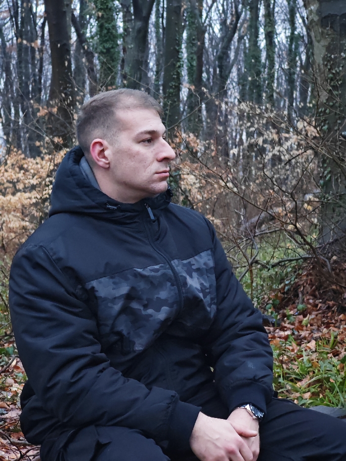 The scene depicts a wooded area in Shumen, Bulgaria, characterized by deciduous trees, likely during late autumn or winter. A single adult male, appearing to be in his 20s or 30s, is positioned in the foreground, seated or crouching, facing right in profile. He has short, light brown hair and a clean-shaven face, exhibiting a neutral expression. The male is dressed in a black hooded winter jacket with a grey and black camouflage pattern across the chest, and black trousers. A silver-toned wristwatch with a dark face is visible on his left wrist, and his hands are clasped together in his lap. The background features numerous bare tree trunks, some partially covered with green ivy. The ground is covered with a layer of brown fallen leaves and sparse green foliage, with dry, light brown underbrush also visible. The ambient lighting is diffused, suggesting an overcast sky.
