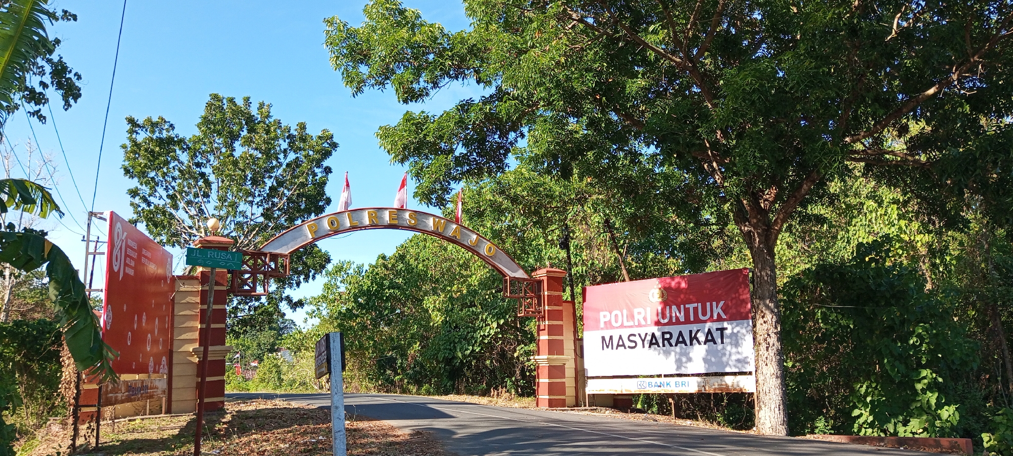 The image captures a bright, sunny day in Bulupabbulu, Indonesia, likely at an entrance to a government facility. Dominating the scene is a large, arched gateway with the words "POLRESWAJO" inscribed in gold lettering across the top. Two Indonesian flags, red and white, are planted atop the arch. Flanking the gateway are tall, red and cream-colored pillars.

To the left of the gateway, a large red banner is mounted. It features a graphic of the number "81" and text in Indonesian, including "INDONESIA". A street sign reading "JL. RUSA I" is attached to one of the gateway pillars.

To the right of the gateway, a prominent red and white banner displays the text "POLRI UNTUK MASYARAKAT" (Police for the Community) in bold white lettering. Below this, a logo for "BANK BRI" is visible.

The surrounding environment is lush and green, with numerous trees casting shadows on the asphalt road leading through the gateway. The sky is a clear, vibrant blue. The overall impression is one of a functional and administrative entrance in a tropical setting.