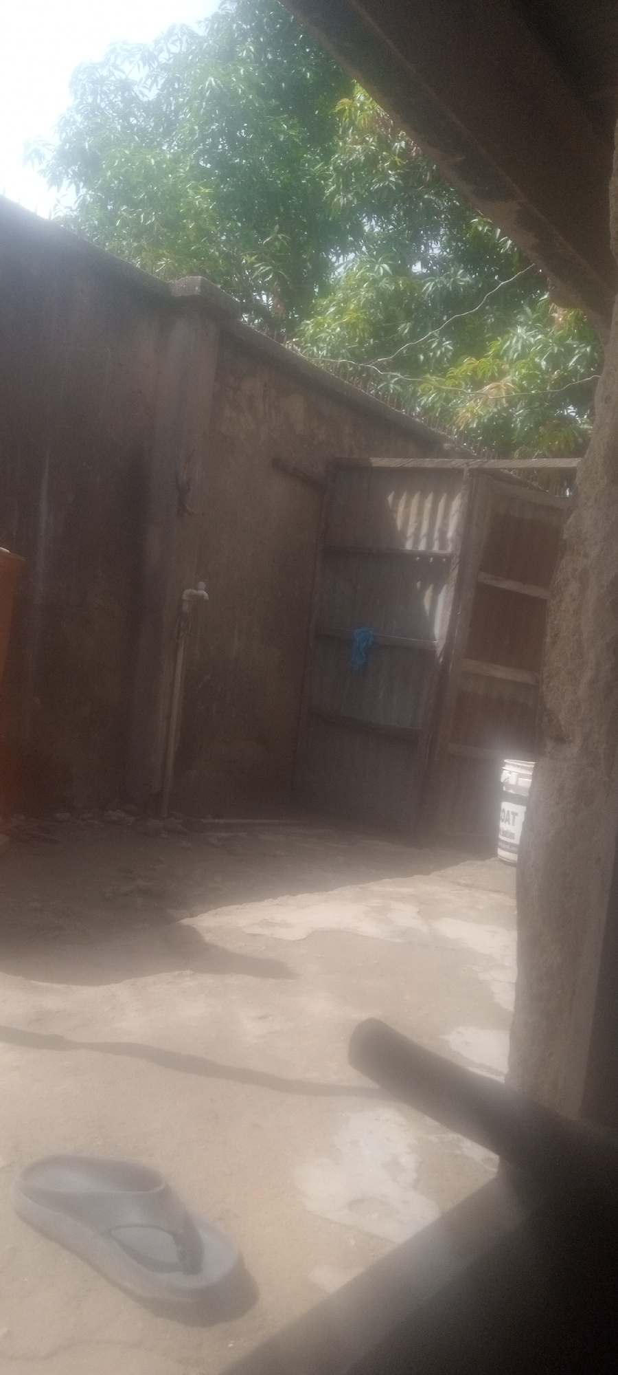 The image depicts an outdoor scene in the city of Unknown, Unknown. A concrete wall with a faucet and a partially open corrugated metal gate are visible. A blue cloth is hanging from the gate. Above the wall, there is vegetation, possibly trees. In the foreground, there is a gray sandal on a sandy surface, and a dark, blurry object partially obscures the lower right corner of the frame. In the background, there is a can of paint, partially obscured. The lighting is bright and somewhat overexposed.