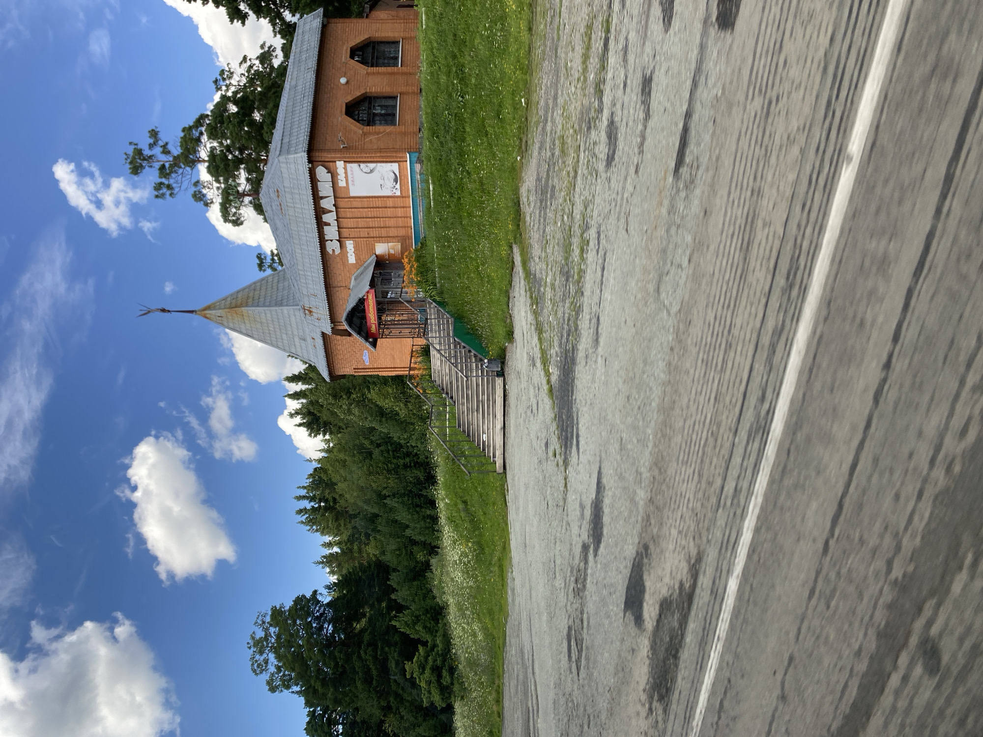 The image captures a brick building with a conical metal roof, situated in the city of Zilair, Russia. The building, which appears to be a cafe or shop, has the word "ЗИЛАИР" (Zilair) prominently displayed on its facade. An awning with the text "Мы работаем!" (We are open!) is visible above the entrance. The building is surrounded by greenery, with a patch of wildflowers on a grassy slope to the left and a dense forest in the background. A paved area, likely a parking lot or road, is in the foreground, with tire marks and scattered shadows indicating the presence of a vehicle from which the photo might have been taken. The sky above is blue with scattered white clouds. No people are visible in the frame.