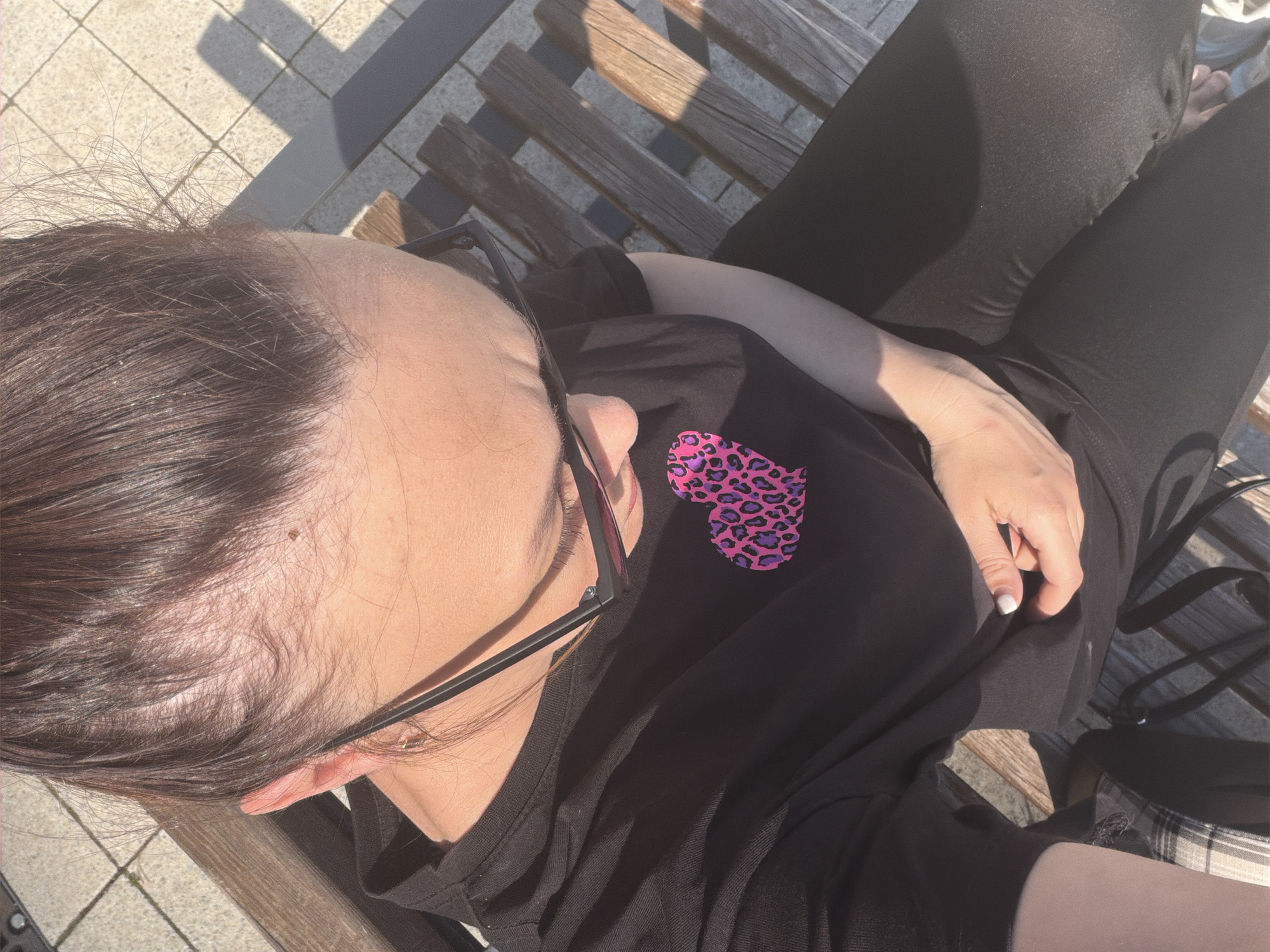 A woman is seated on a wooden bench in an outdoor setting, presumably in the city of Chaikovsky, Russia. The scene is captured from an overhead perspective. The woman is wearing black leggings and a black t-shirt, the latter featuring a pink and purple leopard print heart design. She also has glasses on. Her hair is dark brown. The bench is made of wooden slats with shadows cast upon it. In the background, light-colored paving stones are visible.
