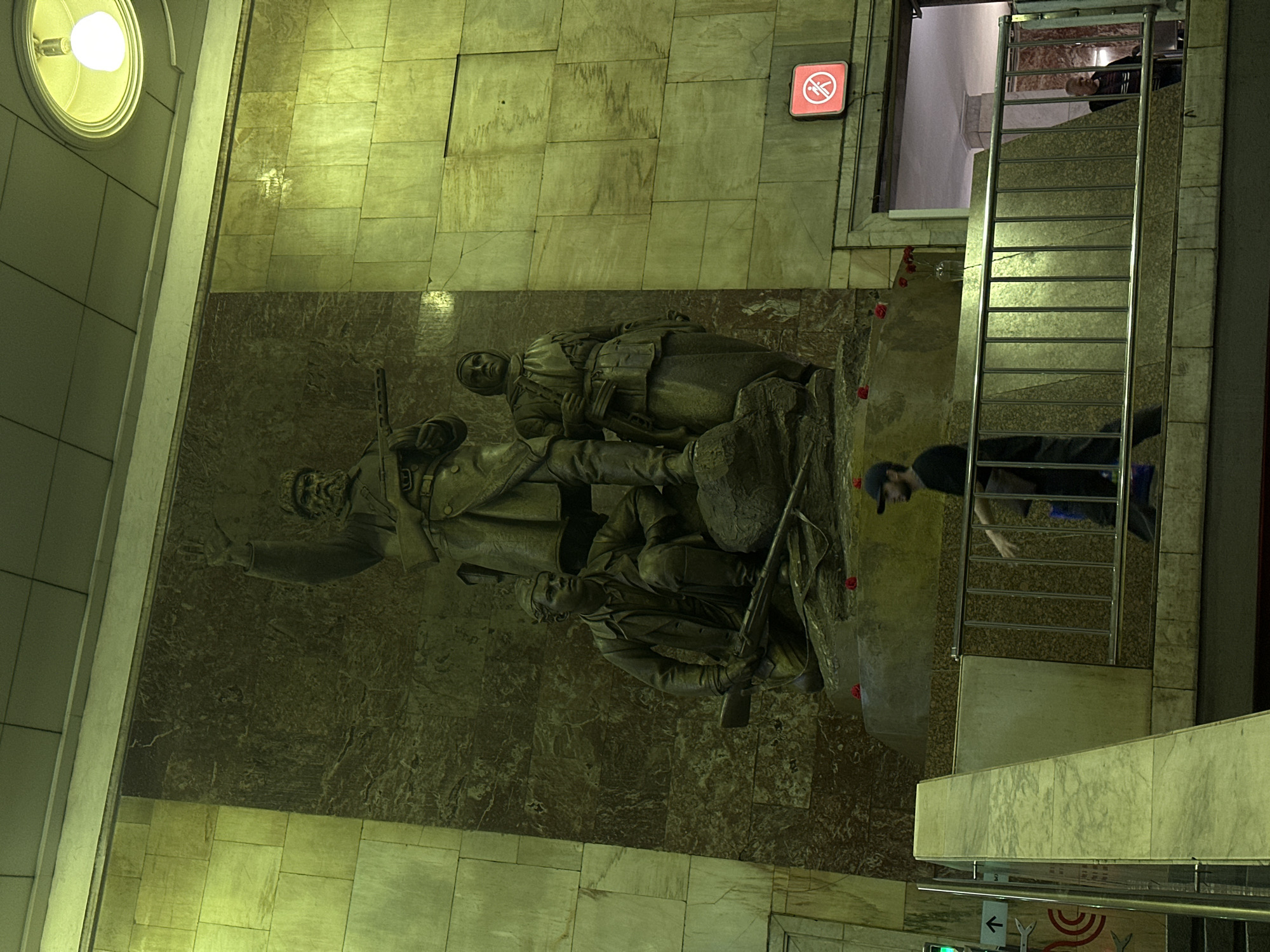This is a medium-wide shot of a large bronze monument located inside a building with marble walls and a tiled ceiling. The monument depicts three figures: a man standing with his arm raised and a rifle, and two figures crouching in front of him, one of whom also appears to be holding a rifle. The figures are dressed in what appears to be military attire. The monument is illuminated by a bright overhead light fixture and stands against a dark marble background.
In the foreground, a metal railing separates the viewer from a ramp or walkway. A man wearing a black t-shirt and a baseball cap is walking along this walkway, with his right arm extended forward. Another person is partially visible behind him. To the right of the monument, a red sign with a white symbol of a person walking is visible above a doorway. Further inside the building, a man in a striped shirt is visible standing in the doorway. The overall lighting in the scene is a yellow-green hue.
This monument is located in the Komsomolskaya metro station in Moscow, Russia, specifically on the Circle Line platform.
