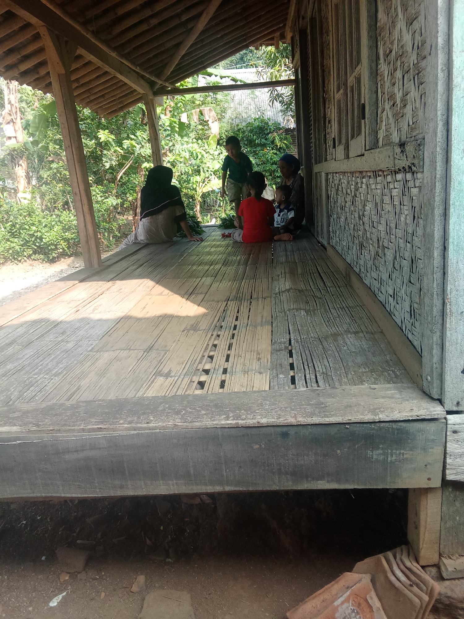 In Cempaka, Indonesia, a group of five individuals, including three children and two adults, are gathered on the wooden porch of a rustic stilt house. The porch floor is constructed from bamboo mats. One adult, wearing a dark headscarf, is seated on the left side of the porch, facing away from the camera. Another adult, wearing a blue headscarf, is seated on the right, also facing away. Three children are present: one child in a red shirt is seated in the center, facing the adults; another child, in a blue and white shirt, is seated next to the child in red. A third child, wearing a dark shirt and shorts, is standing to the left, near the first adult.
The house features walls made of woven bamboo and wooden window frames. The roof of the porch is supported by wooden beams, and the structure is elevated, with the ground beneath visible. Lush green vegetation surrounds the house. Sunlight casts shadows across the bamboo porch floor. In the foreground, under the porch, the ground is visible with some scattered debris and a stack of terracotta tiles. The overall scene depicts a simple, rural domestic setting.
