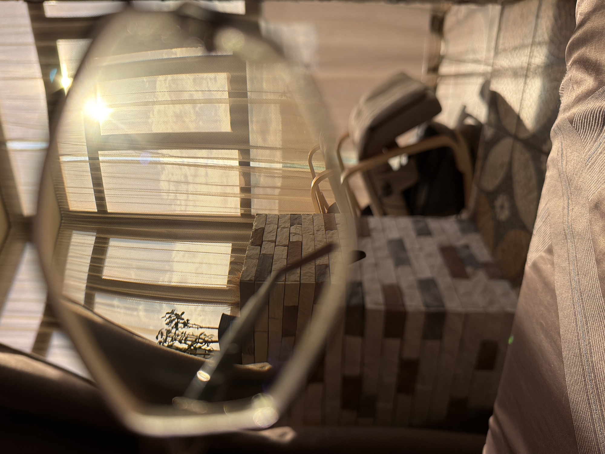 The image depicts an indoor scene, likely a room with a balcony or large window, illuminated by the warm glow of the sun. Through a pair of eyeglasses in the foreground, a view of the room is visible. The room features sheer curtains that diffuse the sunlight. A small potted plant sits on a ledge made of stacked, textured stones. In the background, a rocking chair is positioned on a patterned rug. The overall impression is one of a calm, sunlit interior, potentially a living space. No specific location in Surgut, Russia is identifiable from the visual elements alone.