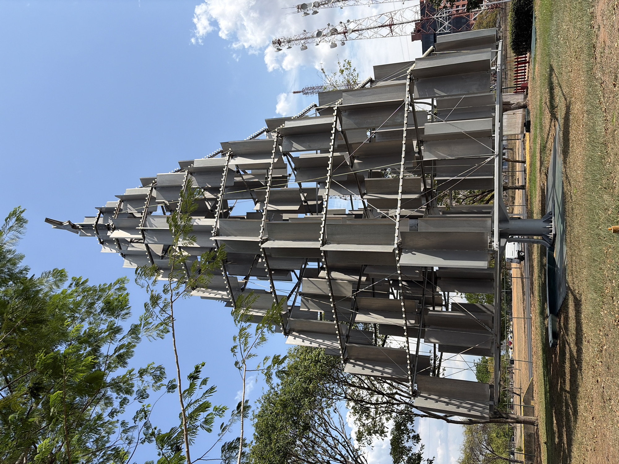 A large, grey, sculptural structure stands prominently outdoors under a bright blue sky with scattered white clouds. The sculpture is composed of numerous horizontal, rectangular panels, likely metal, arranged in staggered layers that taper towards the top, giving it a stylized pyramidal or conical shape. These panels are supported by an intricate metal framework. Small, white, circular elements, possibly lights or decorative studs, are visible in vertical rows along some of the framework's supports.
To the left and partially on the right, lush green trees with distinct foliage frame the sculpture. In the background, on the right side, several tall metal communication towers equipped with various antennas and satellite dishes rise against the sky, alongside a building with exposed structural elements, suggesting construction or industrial activity. The ground is a mix of green and dry, brownish grass, with distinct shadows cast by the structure and trees. A fence runs through the middle ground, separating the immediate grassy area from a more distant, unpaved stretch of ground where faint outlines of vehicles, including a white truck, can be seen.
The scene is captured on a sunny day, evident from the clear sky, bright light, and strong shadows. No people are visible, and no specific event is taking place. There is no discernible text visible in the image. The setting appears to be a public or park-like area within an urban or semi-urban environment, consistent with Ciudad Guayana, Venezuela.