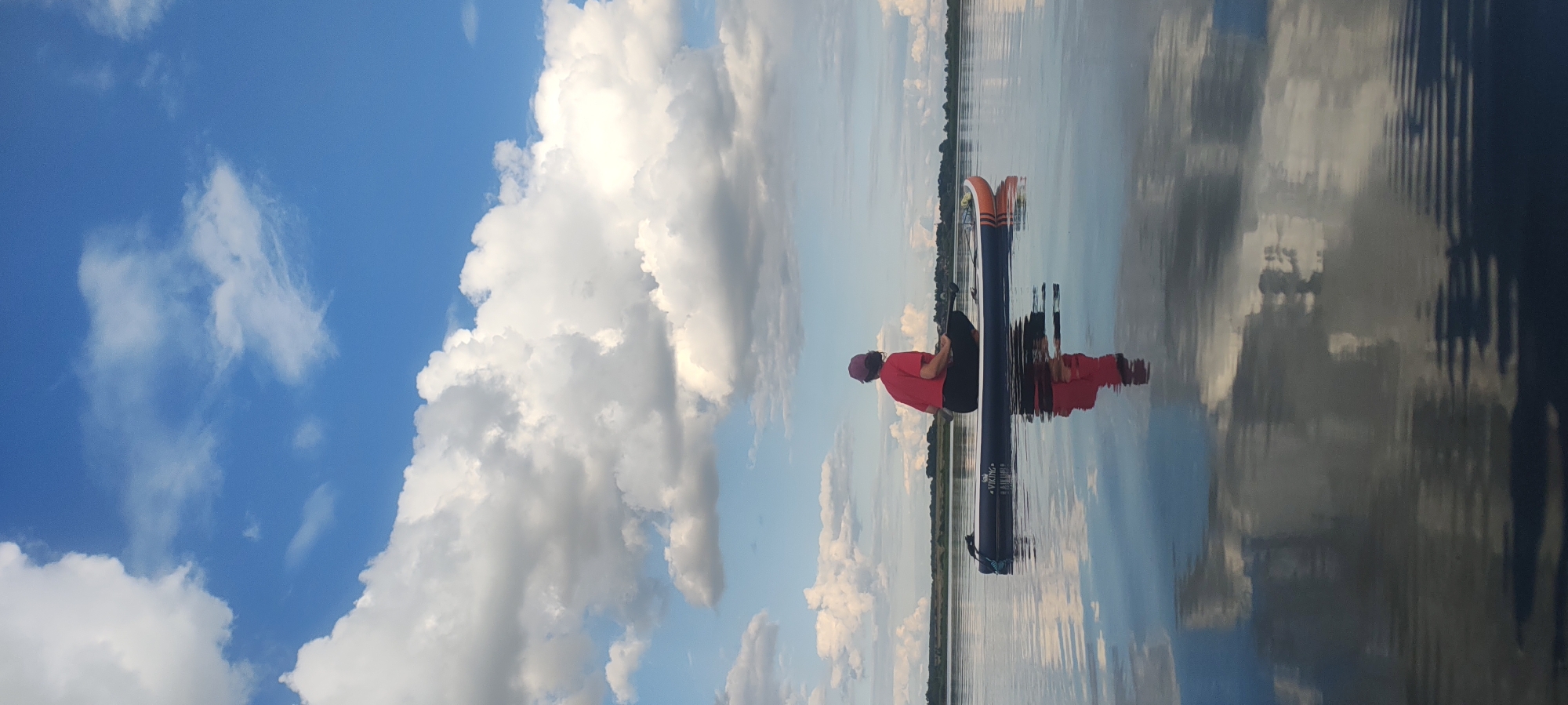 A person is sitting cross-legged on a stand-up paddleboard in the middle of a calm body of water. The person is wearing a red t-shirt and a dark colored hat. They are facing away from the camera, looking towards the horizon. The paddleboard is dark gray with orange and white accents and the brand name "VIKING" is visible. The water is still, reflecting the blue sky with scattered white clouds and the person on the paddleboard. In the distance, a treeline forms the horizon. The scene appears to be in a natural, outdoor environment.