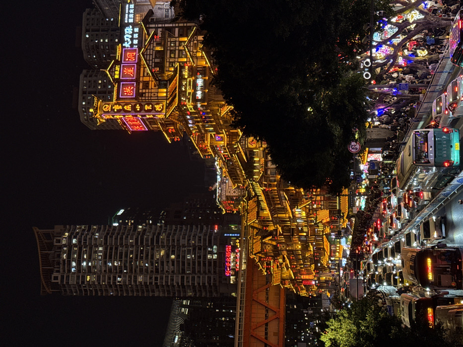 The image displays a nighttime urban scene in Yuzhong District, Chongqing, China. The central feature is a large, multi-tiered complex of traditional Chinese-style buildings, identified as Hongyadong Commercial Street, which ascends a hillside. These buildings are extensively illuminated with golden lights, numerous red lanterns, and vibrant neon signs displaying Chinese characters, including prominent vertical signs at the complex's peak.
In the foreground and midground, a multi-lane asphalt road is heavily congested with vehicular traffic. Numerous white, silver, and dark-colored passenger cars are visible, alongside at least two large buses. Vehicle headlights and taillights are illuminated. A metal barrier separates traffic lanes and pedestrian areas.
To the right of the roadway, a wide pedestrian area is densely populated with numerous individuals walking and congregating. This area is lined with trees featuring dark foliage, some of which are adorned with strings of colorful decorative lights. Streetlights and various traffic signs are also present.
In the background, behind the traditional complex, modern high-rise buildings with illuminated windows are visible against the dark sky. An orange-colored structural bridge or overpass spans the mid-left portion of the image, above another section of illuminated traditional-style buildings. The overall environment is brightly lit and indicates a densely populated, active urban area at night.