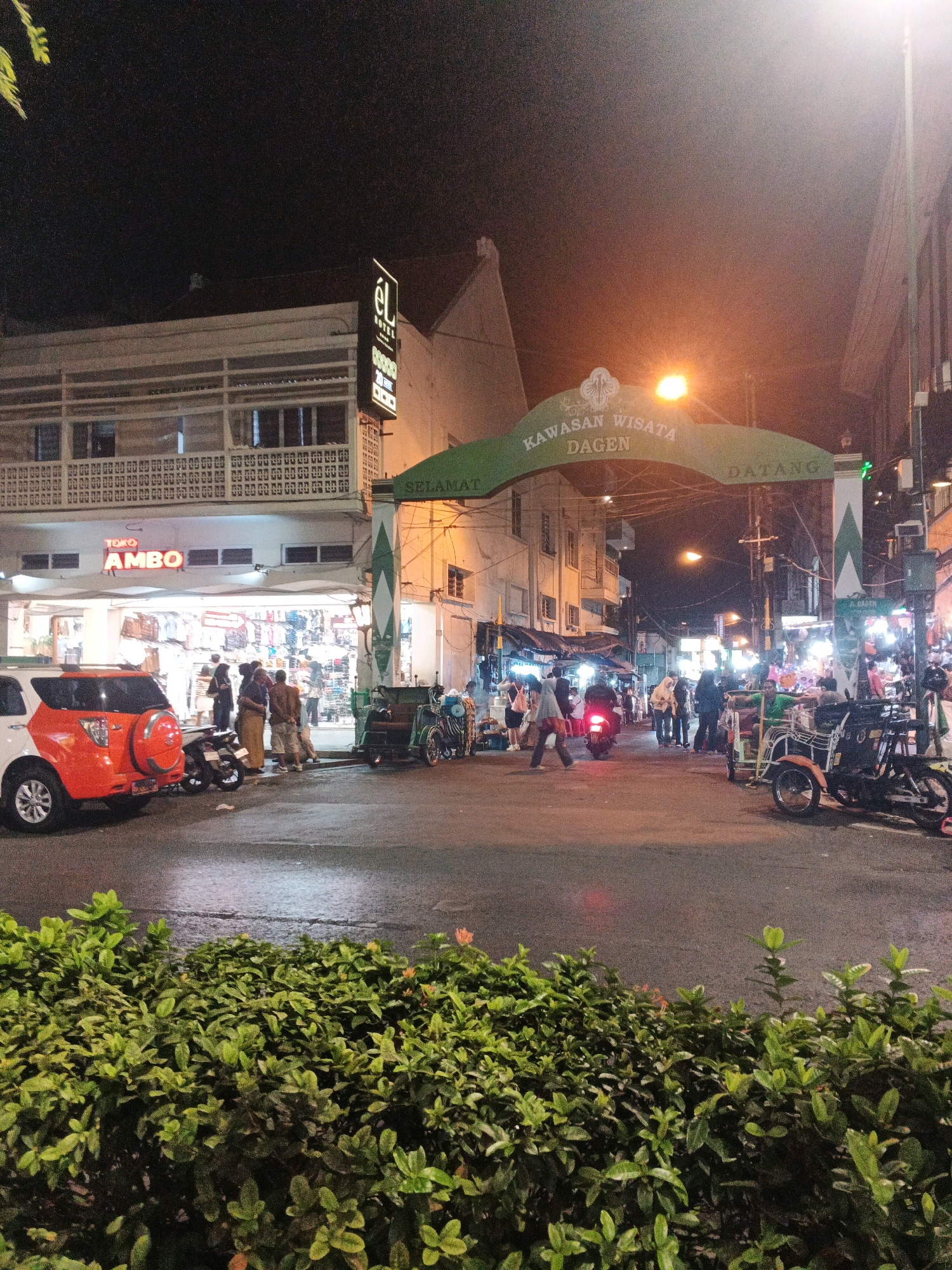 The image depicts a street scene at night in the Malioboro area of Yogyakarta, Indonesia. A large green archway with the text "KAWASAN WISATA DAGEN" and "SELAMAT DATANG" spans across the street, indicating it marks an entrance to a tourist area. Below the archway, a brightly lit store with the sign "TOKO AMBO" is visible, displaying clothing and other merchandise. Several people are gathered outside the store and on the sidewalk.
Vehicles are present on the street, including a white SUV with an orange spare tire cover parked on the left, a motorcycle parked in front of the store, and several traditional Indonesian three-wheeled vehicles (becak) and motorbikes on the right side of the street, some of which appear to be for rent or transport. Streetlights illuminate the scene, creating reflections on the wet asphalt. Dense green foliage is in the foreground, partially obscuring the view of the street. The overall atmosphere is that of a bustling night market or shopping district.