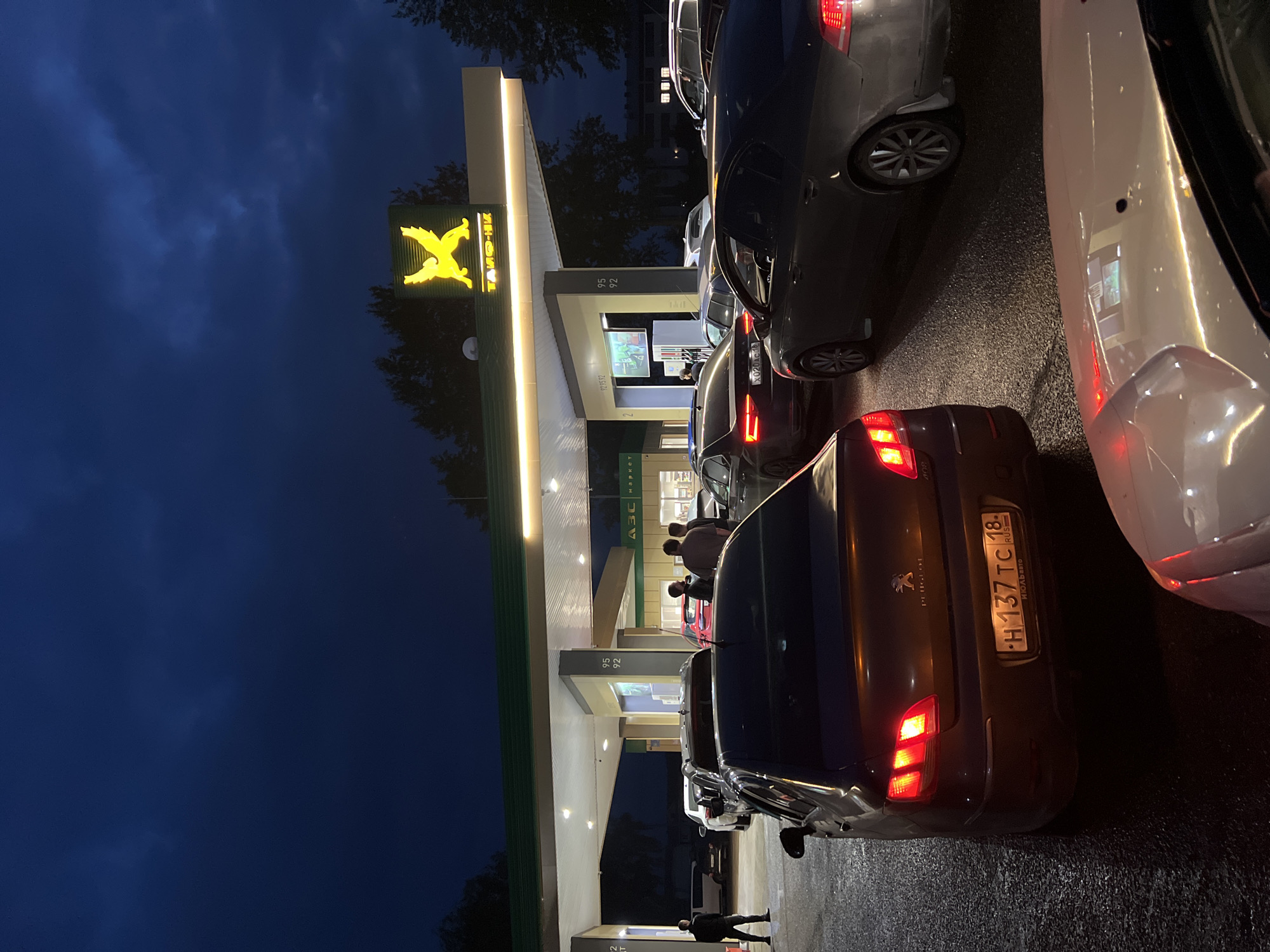 This media file captures a nighttime scene at a Tamneft gas station in Agryz, Russia. The image shows a queue of cars lined up at the pumps, suggesting a potential shortage or increased demand for fuel. The gas station canopy is illuminated by fluorescent lights, casting a glow on the wet asphalt below, indicating recent rain or damp conditions.
The prominent yellow logo of Tamneft, featuring a stylized bird, is clearly visible on the side of the station. Next to the logo, the name "TAMQ-H" is also visible in Cyrillic script. The pumps themselves display fuel grades, with "95" and "92" clearly legible. A digital display on one of the pumps shows "5522".
Several cars are visible in the queue. In the foreground, a dark-colored Peugeot with the license plate "H 137 TC 18" is parked. The taillights of this car are illuminated. Other vehicles, including a white car and a dark gray car, are also part of the line. Some individuals are seen standing near the pumps, possibly refueling their vehicles or waiting their turn. The overall mood is one of quiet activity under the artificial lights of the gas station, with the dark sky indicating late evening or night. The presence of multiple cars suggests a common, everyday event of refueling, but the queue might hint at a more urgent need or situation.