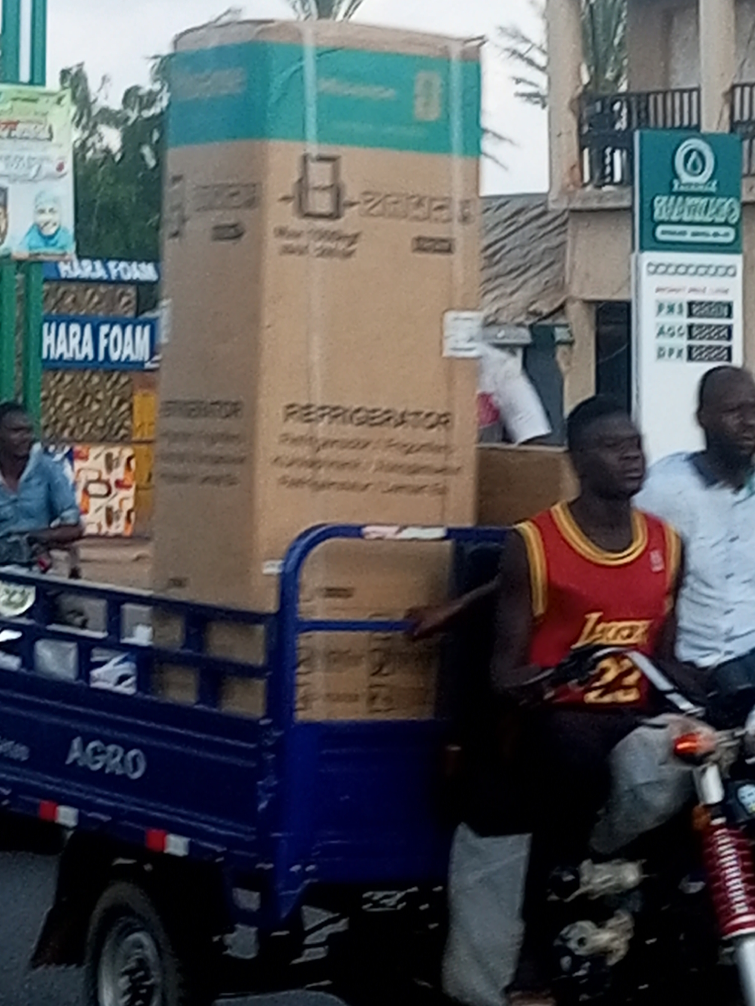 The image shows a flatbed truck loaded with large cardboard boxes, likely appliances, in Garko, Nigeria. The truck is blue and has the word "AGRO" written on its side. Two men are visible on the truck, one in the foreground wearing a red basketball jersey with the number 23 and the Lakers logo, and another man in the background wearing a light blue shirt. There are other people visible in the background, some looking towards the truck.
On the left side of the image, there are signs that say "HARA FOAM" and a picture of a child. In the background, there is a building with a sign that appears to be for a gas station, with prices for "PMS", "AGO", and "DPX" visible. The overall scene suggests a commercial area in Nigeria, possibly during the daytime given the lighting, though the weather is unclear. The men appear to be engaged in or observing the loading or unloading of goods.