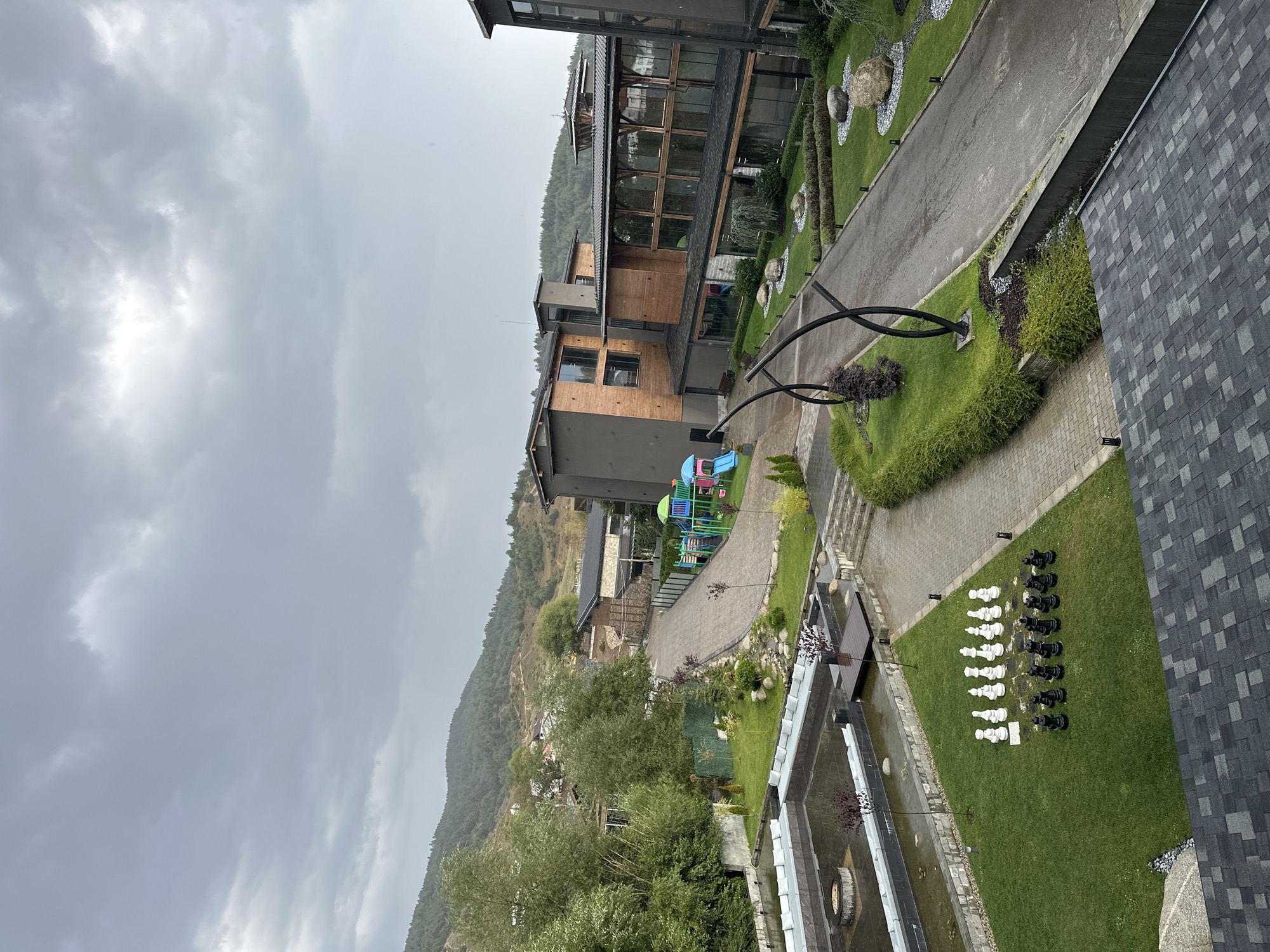 This image captures an outdoor scene at a resort in the city of Bania, Bulgaria. In the foreground, a dark grey asphalt roof with a checkerboard pattern of shingles is visible. To the left, a rectangular water feature with an outdoor fire pit and L-shaped seating arrangements surrounds it. Adjacent to the water feature is a manicured green lawn featuring a life-sized chess set with black and white pieces.
Beyond the lawn, a paved walkway leads to a playground equipped with colorful slides and climbing structures. Further back, modern, multi-story buildings with wooden and dark grey facades dominate the landscape. Large windows on these buildings reflect the overcast sky. A paved road curves through the property, bordered by green lawns, decorative rocks, and sculpted bushes. In the background, a lush, forested mountain range rises under a cloudy sky, with a few houses scattered on its slopes. The overall atmosphere suggests a serene, contemporary resort setting amidst natural beauty.