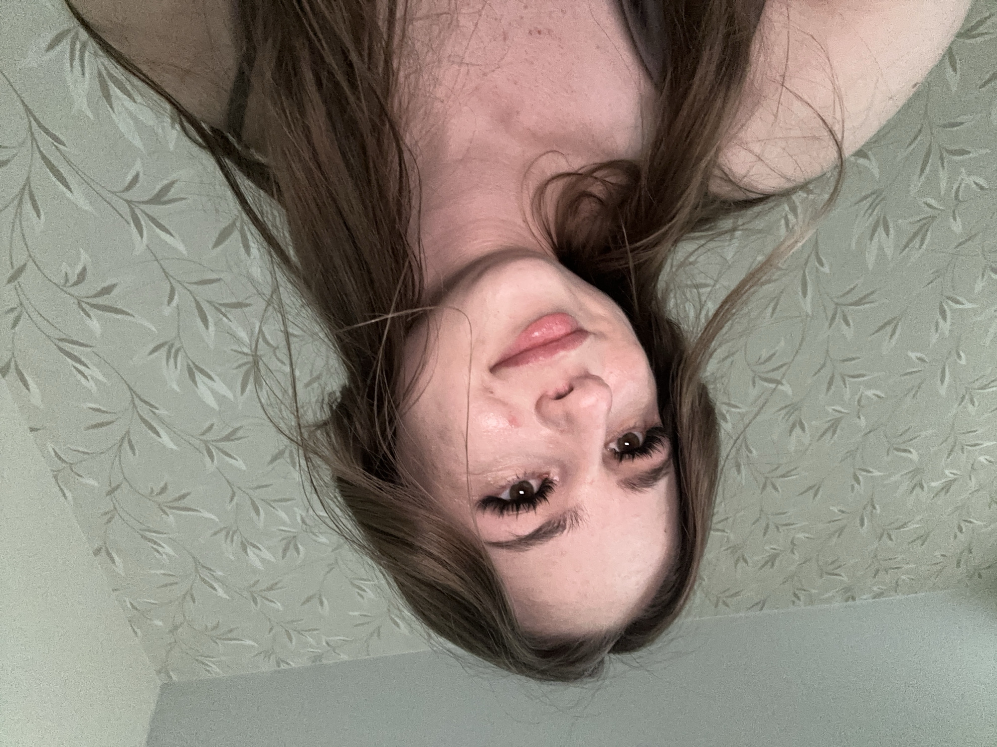 The media file is a close-up, upside-down selfie of a young woman with long, brown hair. She has light skin, prominent eyelashes, and is wearing light makeup. Her expression is neutral. The background appears to be a wall with a light-colored floral wallpaper. The lighting suggests it may be indoors during the daytime. There is no visible text in the image.