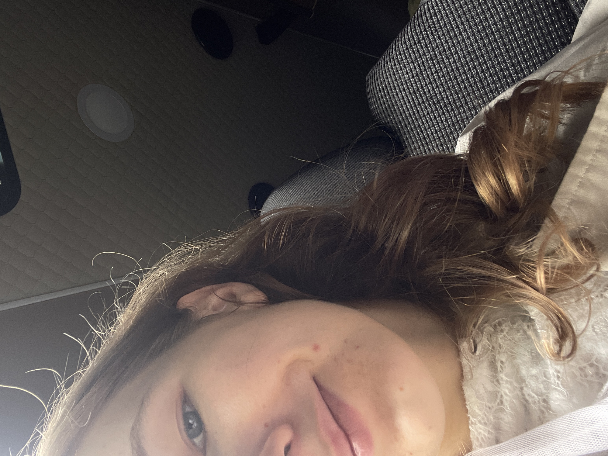 An unidentifiable person's face is partially visible on the left side of the frame. The individual has light skin with a few blemishes, blue eyes, and light brown hair. They are wearing a white, textured garment. The background appears to be the interior of a vehicle, with a quilted ceiling panel, a circular light fixture, and patterned seating visible on the right. The scene is lit from above and to the left, casting shadows on the right side of the frame. The photograph was taken in Ulmen, Germany.
