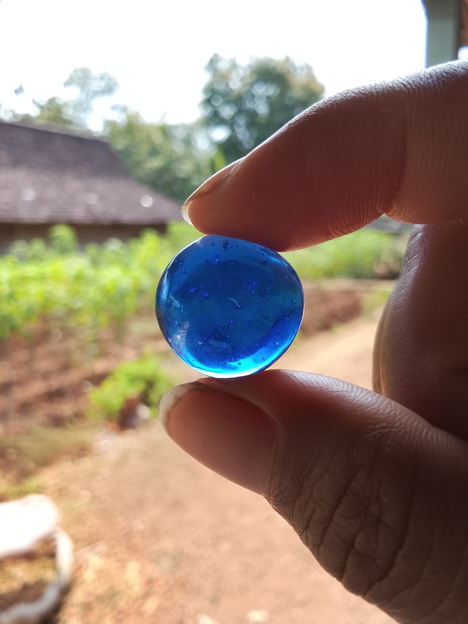 An extreme close-up shows a person's thumb and index finger holding a translucent blue glass marble. The marble has small air bubbles within it. The background is blurred, showing a house with a brown roof and lush green foliage, suggesting an outdoor environment. The ground in the background appears to be dirt or dry earth. The scene is set in the city of Dawung, Indonesia.