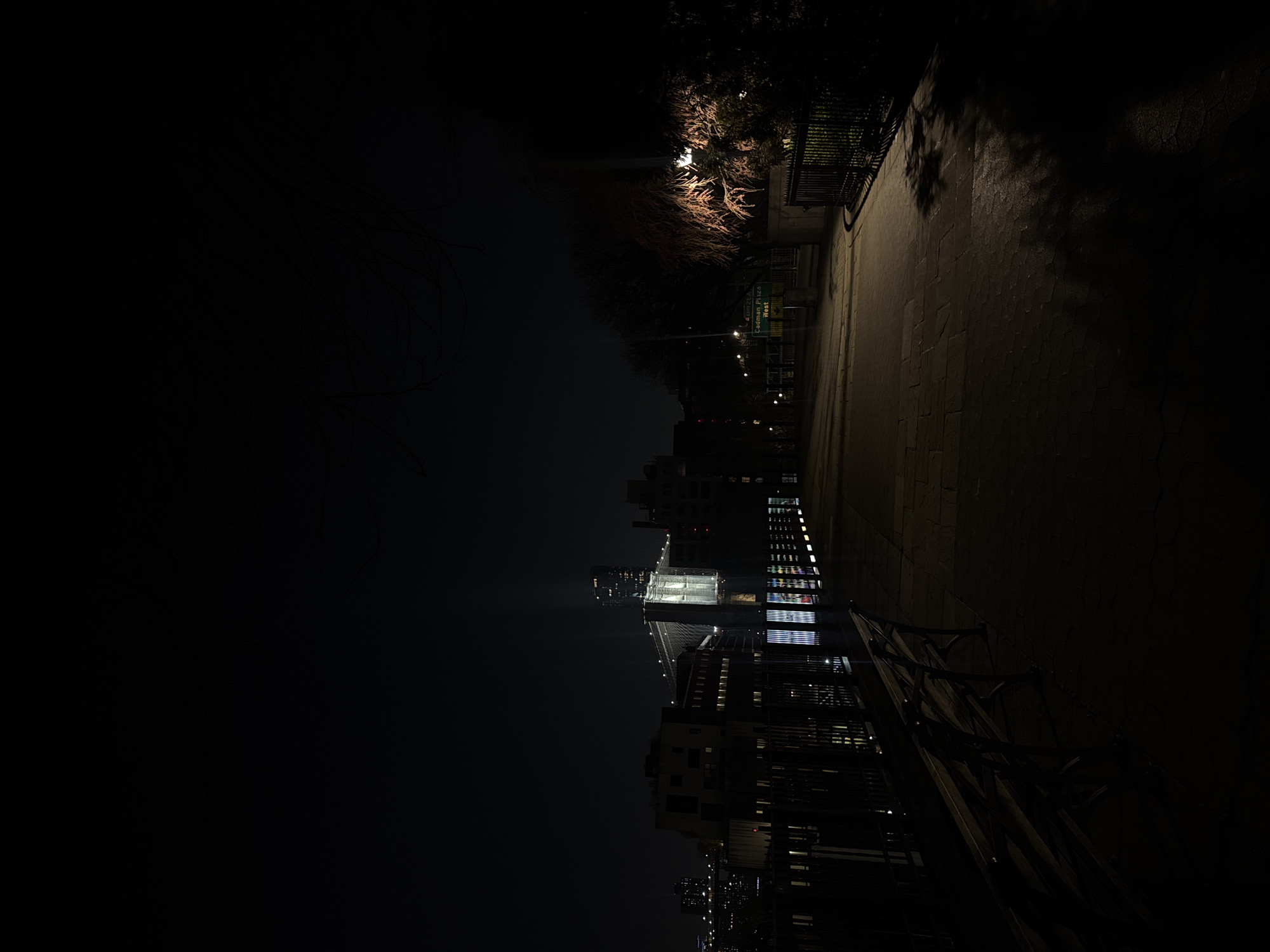 The image depicts a night scene in New York City. On the right side of the frame, a dimly lit sidewalk stretches into the distance, bordered by a fence and some foliage. The left side is dominated by darkness, gradually revealing a cityscape skyline with illuminated buildings. A bridge is prominently featured, lit up against the night sky. The overall ambiance is dark and urban.