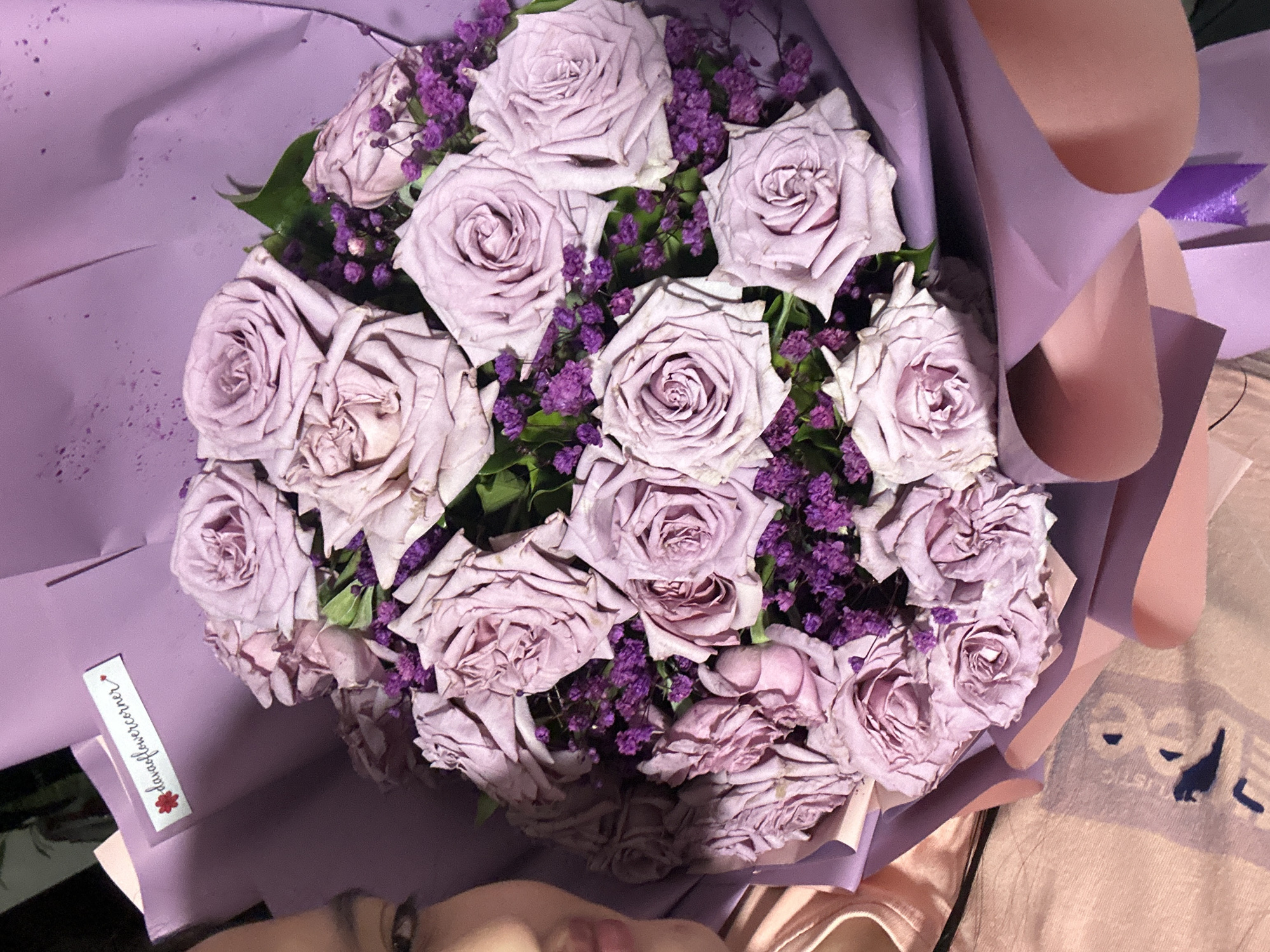 The image shows a bouquet of lavender roses with small purple filler flowers, wrapped in lilac paper. A small rectangular label with "davaoflowercorner" and a flower graphic is visible on the wrapping. The bouquet appears to be resting on a surface, possibly a lap, with a partial view of a person's face and clothing in the lower part of the frame. The location mentioned in the description is Davao City, Philippines.