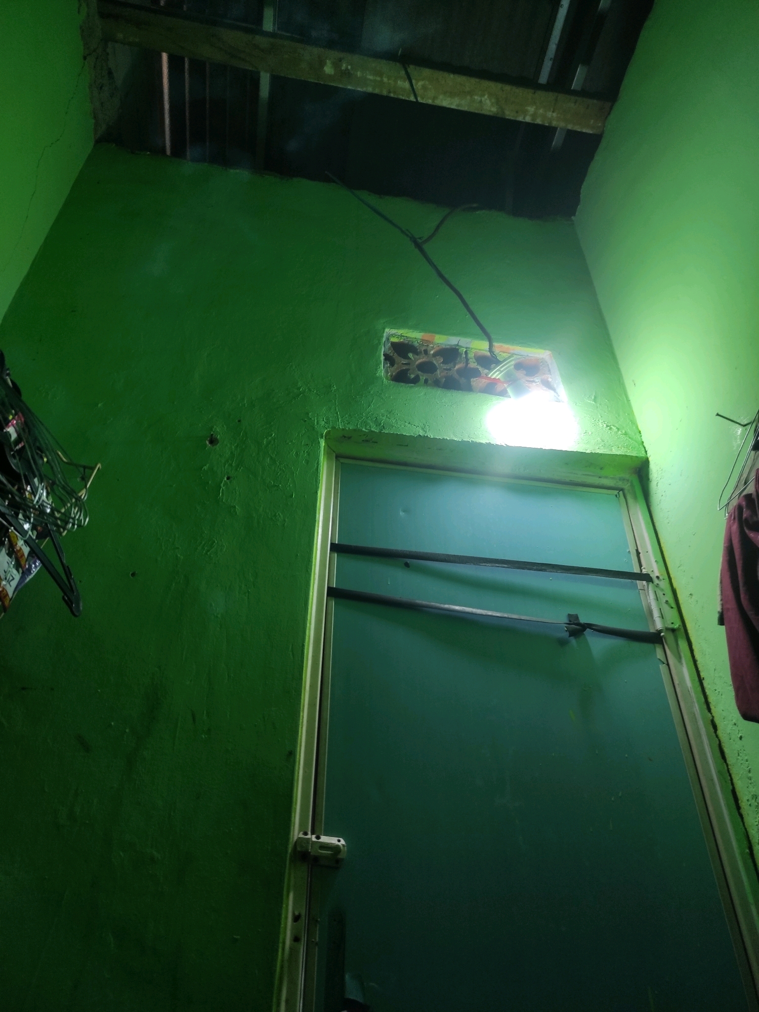 The image depicts a dimly lit interior space with walls painted a vibrant green. In the upper part of the image, exposed wooden beams and metal framing suggest an unfinished ceiling or roof structure. A bare light bulb hangs from a wire, casting a bright light and illuminating a section of the wall above a doorway. The doorway itself appears to be a light blue or teal color, with two horizontal metal bars across its upper half. To the left of the doorway, a collection of dark-colored items, possibly hangers or clothing on hangers, is visible. A small, rectangular window with a decorative grille is situated above the doorway. On the right side of the doorway, a piece of maroon fabric hangs against the green wall. The overall impression is of a humble, possibly utilitarian space. This scene is located in the city of Padalarang, Indonesia.