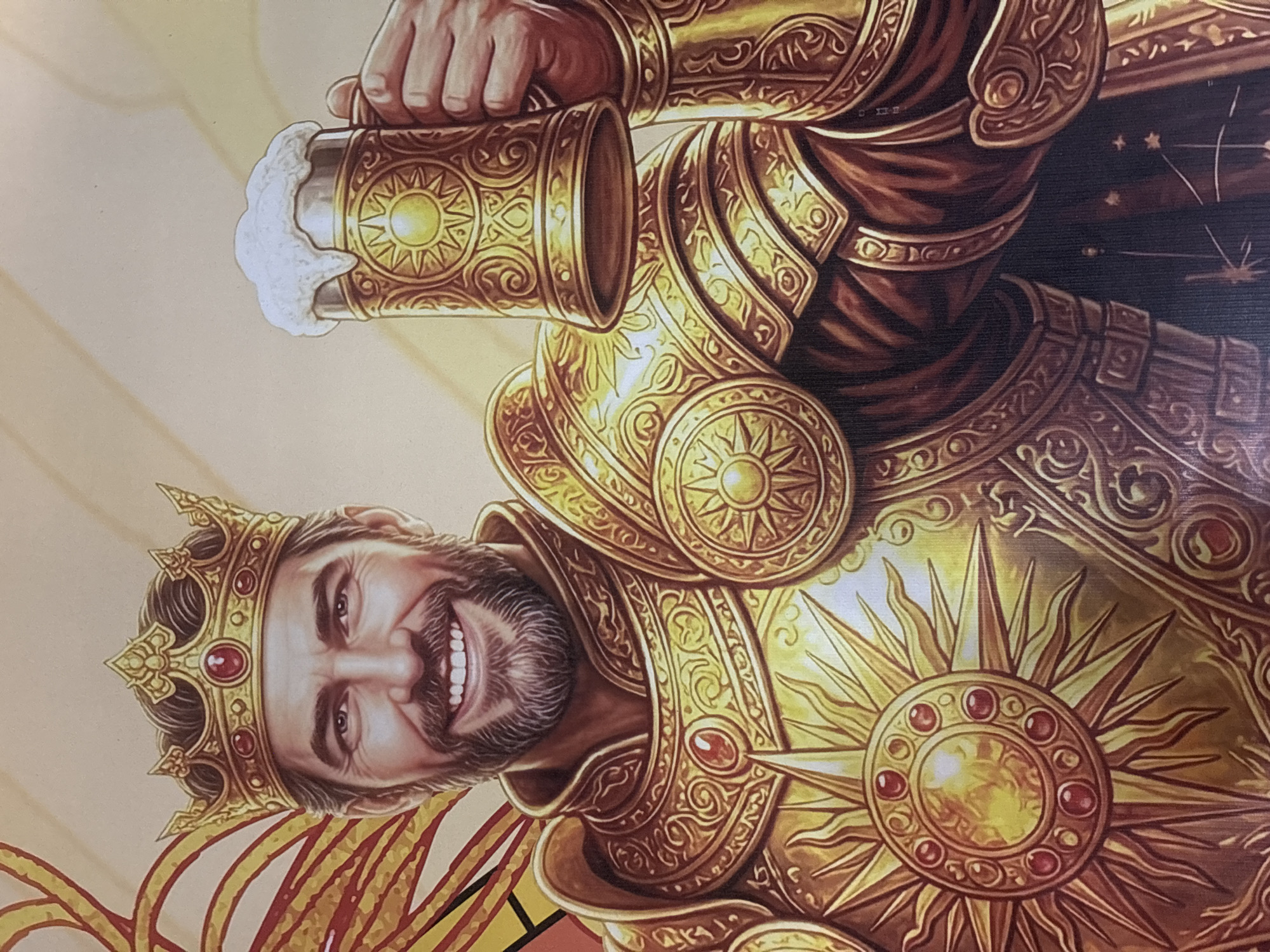The image depicts a richly detailed illustration of a man wearing a golden crown and ornate golden armor. He has a beard and is smiling, holding a frothy mug of beer. The armor is heavily decorated with sun motifs and red gemstones. The background is a warm, gradient of yellow and beige, with some red and yellow ornamental elements on the left side. The image appears to be a digital painting or illustration, not a photograph of a real-world scene. There is no indication of any specific location in Đà Nẵng, Vietnam, or any other geographical location.