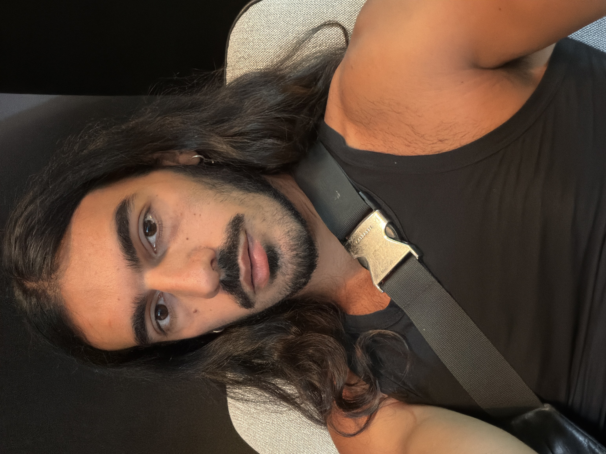 A close-up, diagonal portrait features an adult individual with long, dark, wavy hair, a full beard, and a mustache. The subject has dark eyes and prominent eyebrows, looking directly into the camera with a neutral to slightly serious expression. They are wearing a dark, possibly black, sleeveless top or tank top. A dark gray strap, resembling a seatbelt or a bag strap, runs diagonally across their chest, secured by a silver metallic buckle engraved with the text "1017 ALYX 9SM". A silver hoop earring is visible in the subject's left ear.
The person appears to be reclining, with their head resting against a light gray, textured upholstered surface, suggesting a seat or cushion. The background above their head is dark and indistinct. Body hair is visible on the subject's arm and shoulder. The lighting is soft and even, without specific indicators of time of day or weather. While the context suggests Kuta, Indonesia, there are no visual elements within the image to confirm this location. The overall scene conveys a casual and relaxed posture.