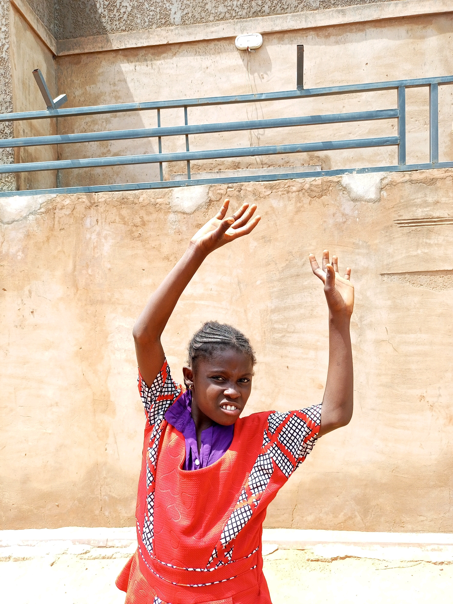 A young Black girl, appearing to be in her pre-teen years, stands outdoors in front of a textured, light-brown wall. She has braided hair and is wearing a bright red dress with a black and white geometric pattern on the sleeves and trim, along with a purple collar. Her arms are raised, with her hands open, as if in a dance or gesture of celebration. She has a slight grin on her face. Behind her, a metal railing runs horizontally across the wall, and a textured concrete structure is visible at the top of the frame. The ground in front of her appears to be dirt or sand.