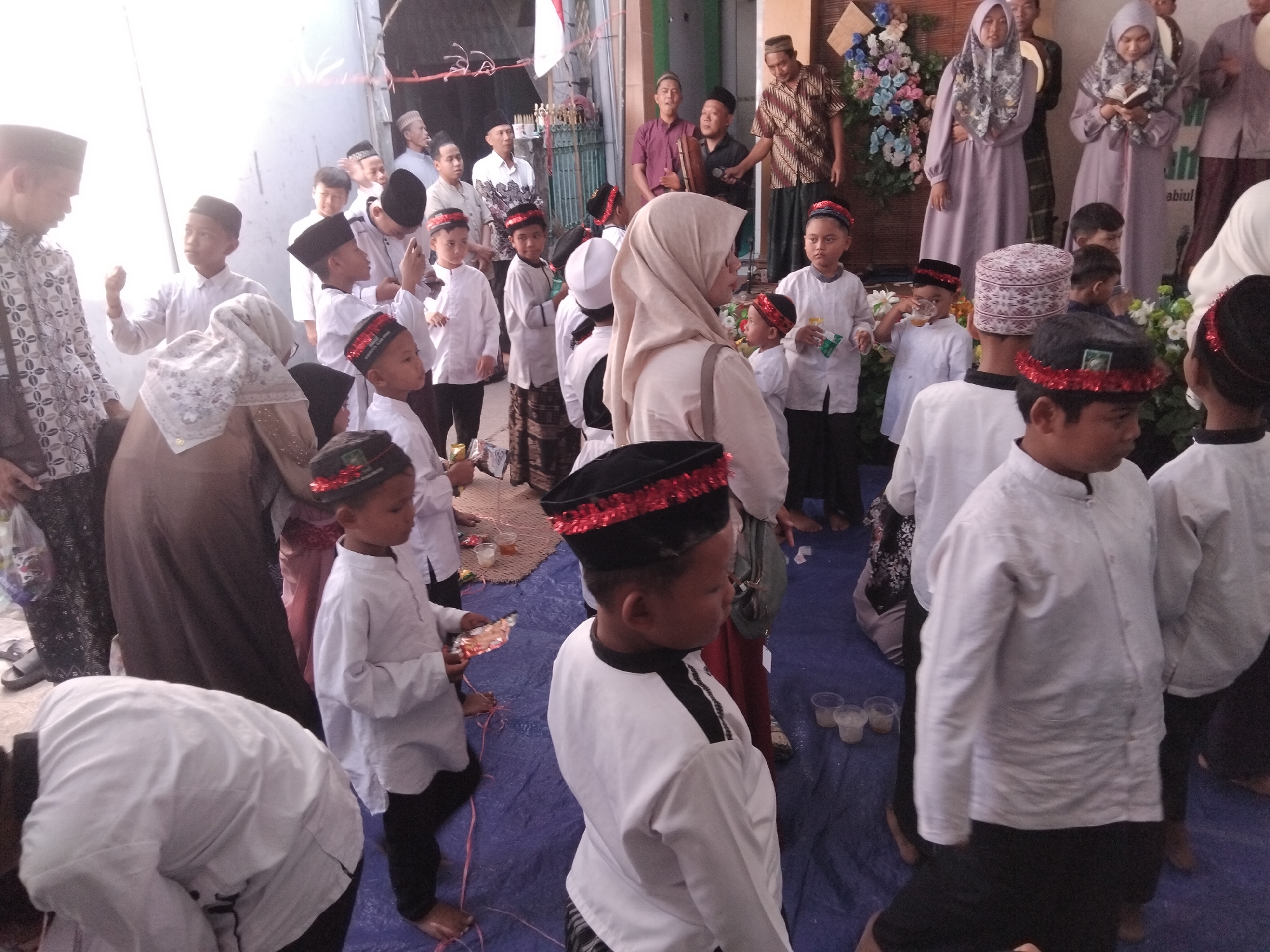 The image depicts a gathering of people, primarily children and adults, in an outdoor or semi-outdoor setting that appears to be a community event. Many of the children are wearing white shirts and black pants, some with black caps adorned with red trim. Several adults are also present, some wearing traditional Indonesian attire. The background includes a structure that could be a building or tent, adorned with a floral arrangement. The ground is covered with a blue tarp, and there are some woven mats and plastic cups visible, suggesting refreshments or a meal. The overall atmosphere suggests a religious or cultural celebration. The image is identified as being from Kepuh Kiriman, Indonesia.