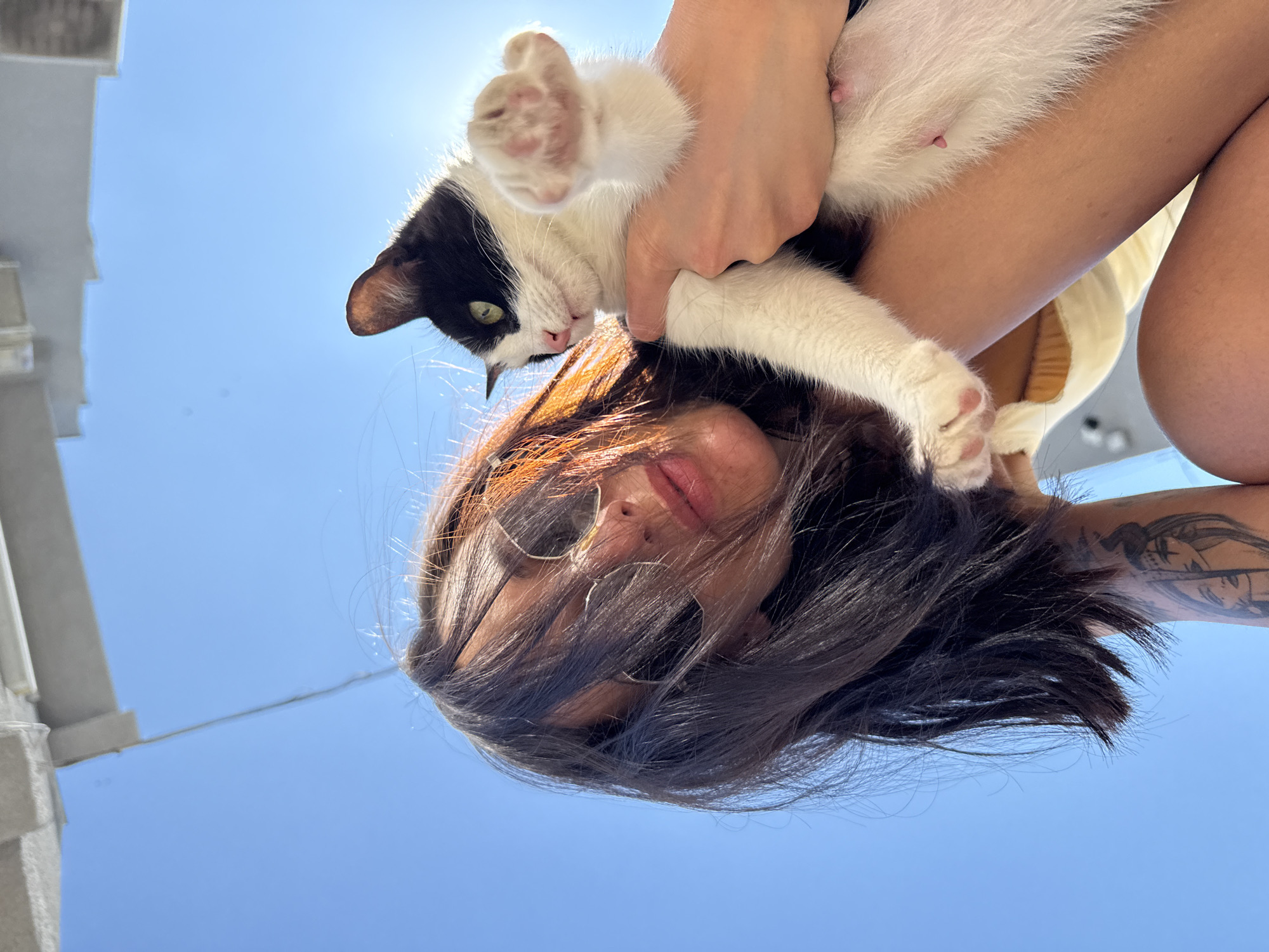 A low-angle shot features a woman holding a black and white cat against a clear, blue sky. The woman has dark hair, sunglasses perched on her face, and a tattoo visible on her arm. She is holding the cat, which is facing the camera, with one paw extended. The cat is a black and white female and has visible nipples. The photograph appears to have been taken outside, with a partial view of a building visible in the background. There is no information to confirm the location of this photo is Pleven, Bulgaria.