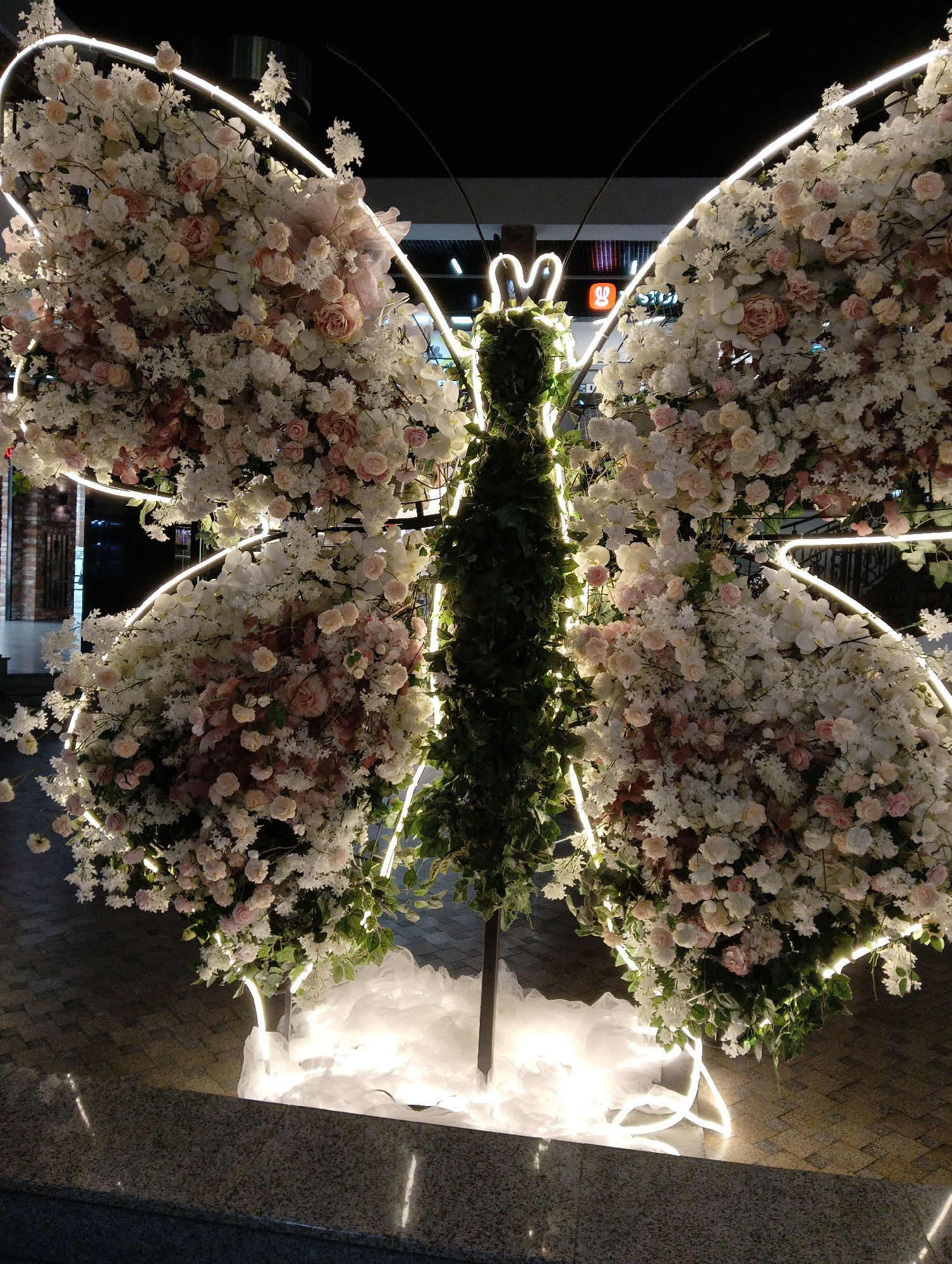 This is a full-frame, eye-level, outdoor, night-time shot of a large, illuminated butterfly-shaped floral display. The display is composed of artificial white and pink flowers, with accents of green foliage in the center that forms the body of the butterfly. White neon lights outline the wings and antennae. The display is situated on a patterned paved surface, with a granite-like border in the foreground. In the background, glimpses of a building with illuminated signage and windows can be seen. There are no people or animals visible in the image. The overall impression is one of a decorative, possibly festive, installation. The city of Hrodna, Belarus, is not visually identifiable from the provided image.