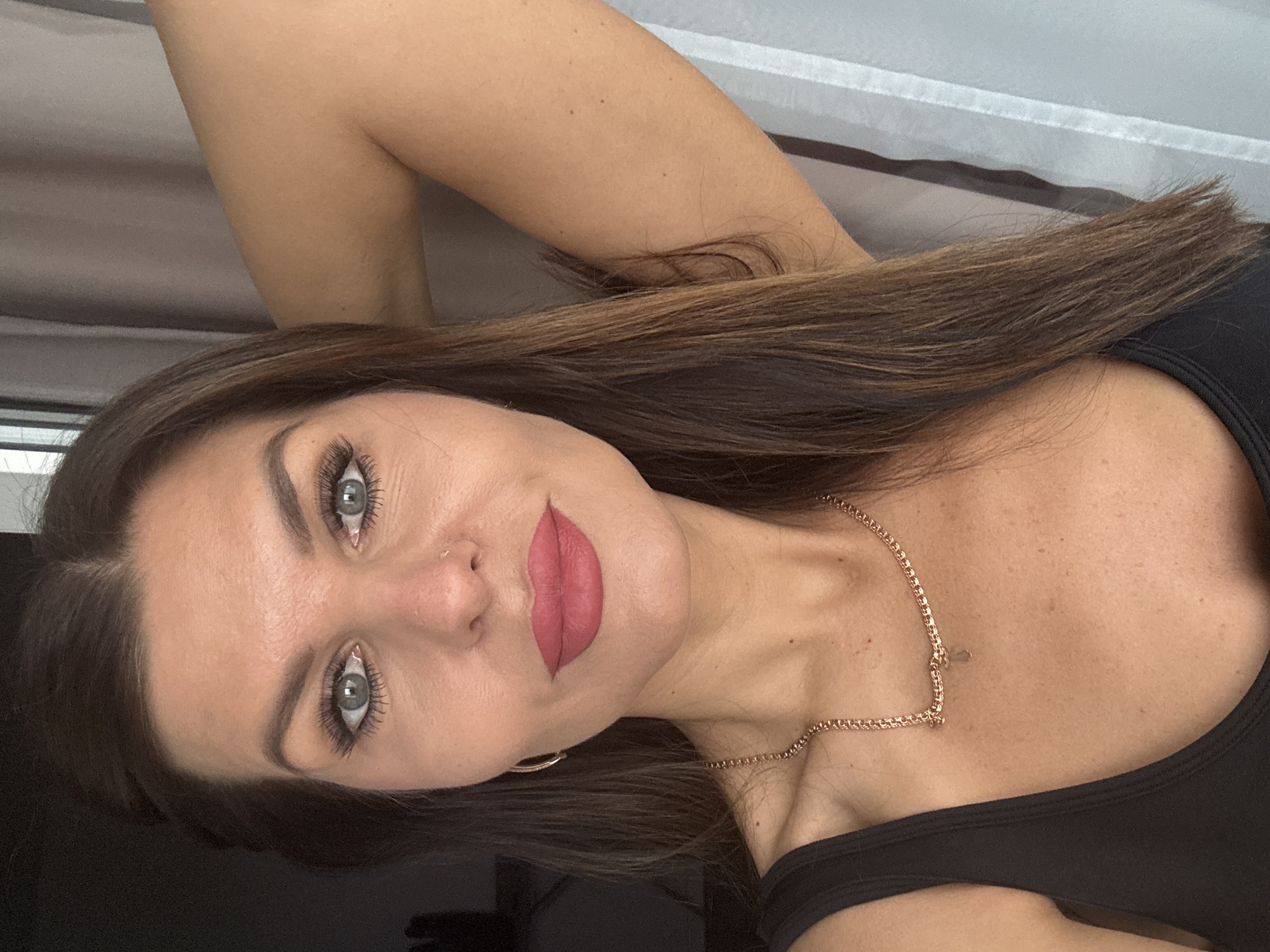 The image is a medium close-up portrait of a woman, oriented 90 degrees counter-clockwise. The woman's head is on the left side of the frame, and her body extends towards the right. She has long, dark brown hair flowing around her shoulders and arms. Her striking eyes appear green-grey or light blue, accentuated by prominent dark mascara on her eyelashes and well-groomed, dark eyebrows. Her lips are painted with a soft, matte red or deep pink lipstick, and are slightly parted.
She wears a dark-colored (likely black) tank top or camisole. A delicate, gold-colored chain necklace is visible around her neck, draped over her collarbone. Her expression is calm and neutral. The background is softly blurred, suggesting an indoor setting with lighter tones, possibly a wall or curtain, in the upper right portion of the frame. The image captures a posed moment, likely a casual portrait. There is no visible text in the image. This photograph was taken in Chaikovsky, Russia.