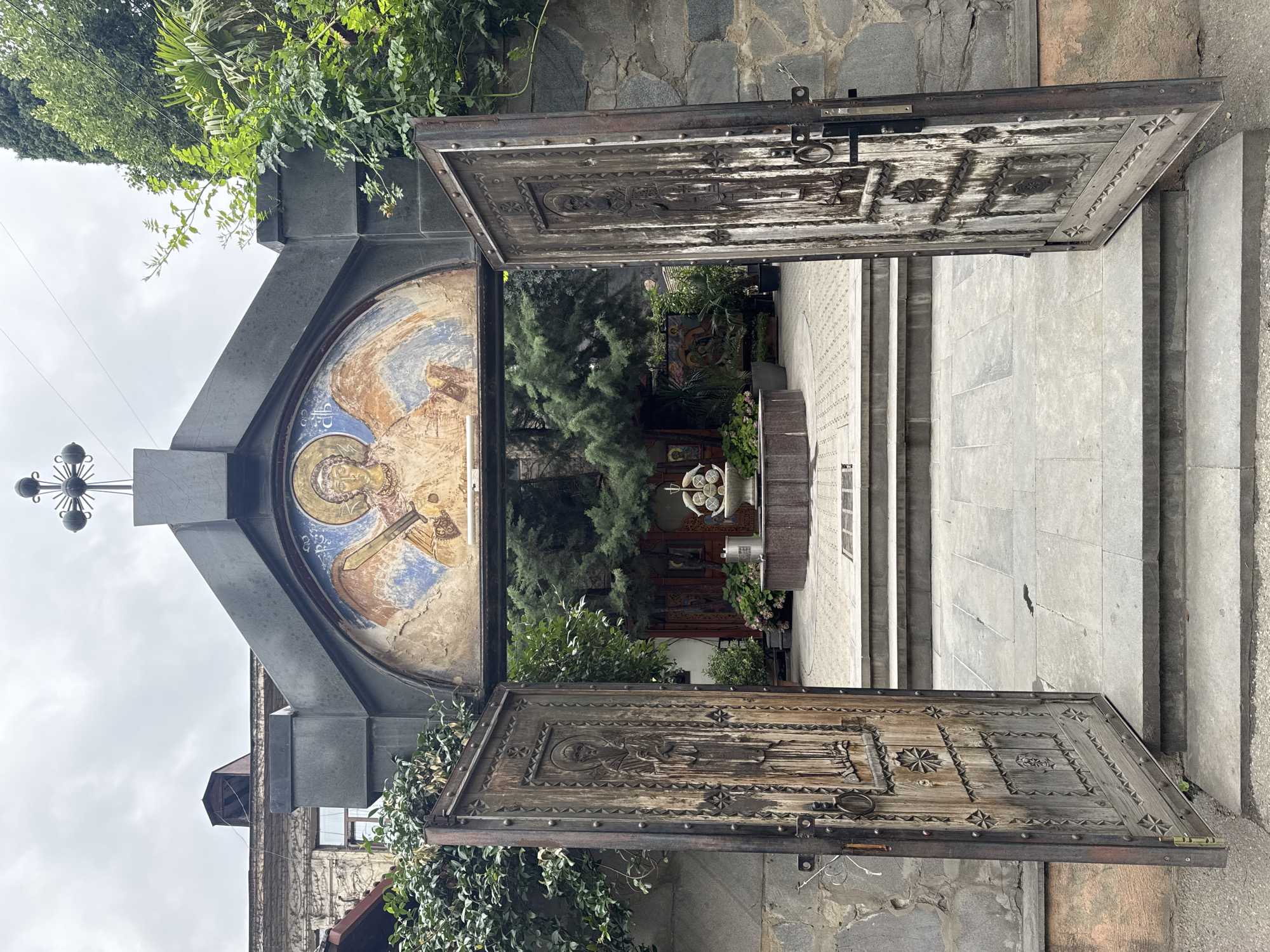 The image shows an open set of weathered wooden double doors leading into a courtyard in Tbilisi, Georgia. Above the entrance, an arched mosaic depicts an angel with a sword. The courtyard contains a fountain, greenery, and religious icons. The doors are heavily ornamented with carved figures and metal studs, showing signs of age and wear. The architecture suggests a historic church or monastery.
