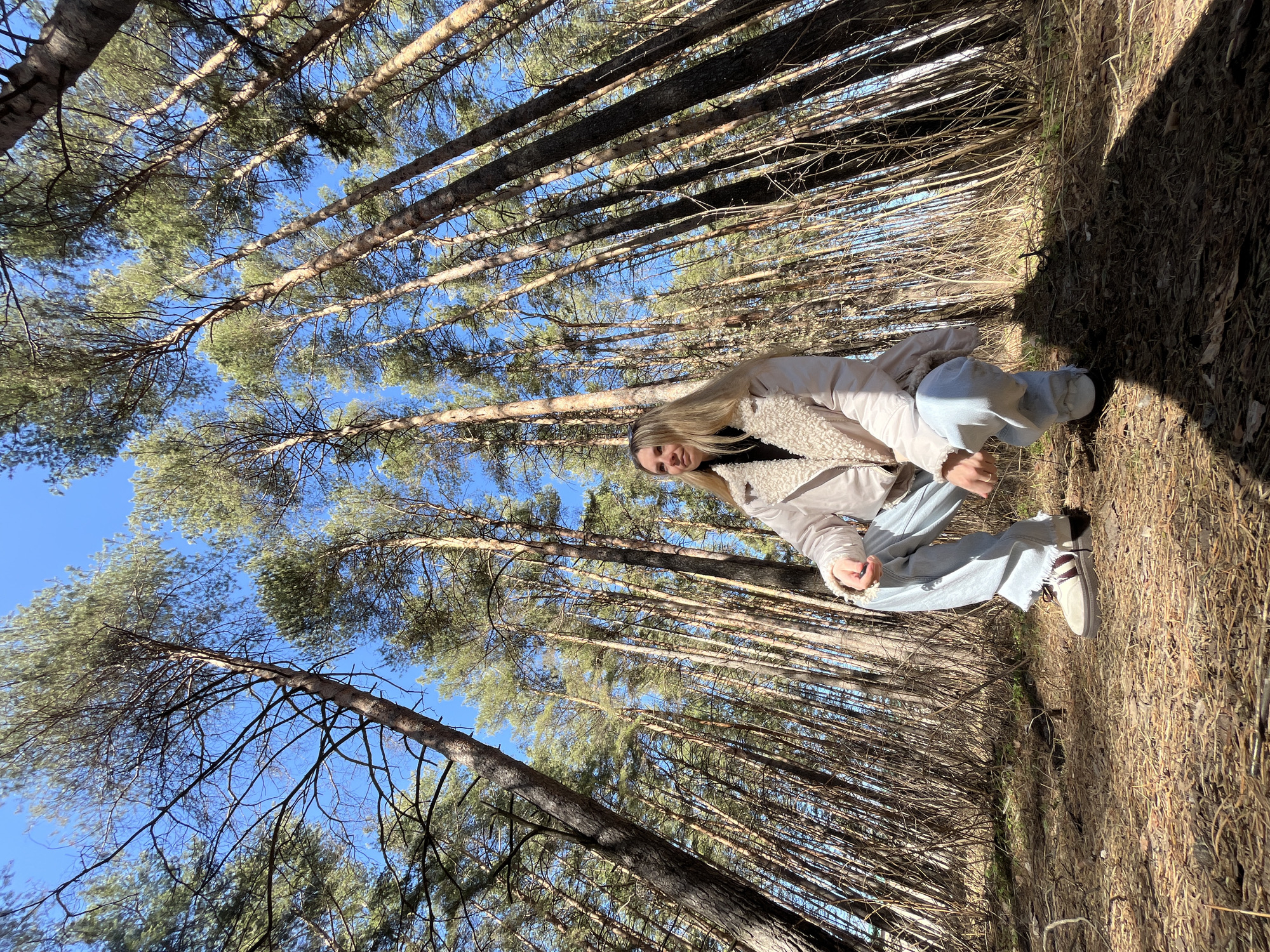 A woman is squatting in a pine forest. She has long, blond hair and is wearing a light-colored jacket, black shirt, and light blue jeans. She's looking directly at the camera. The scene is set among tall, straight pine trees with a clear blue sky visible through the canopy. The ground is covered with dry leaves and pine needles. The location is the city of Chaikovsky, Russia.