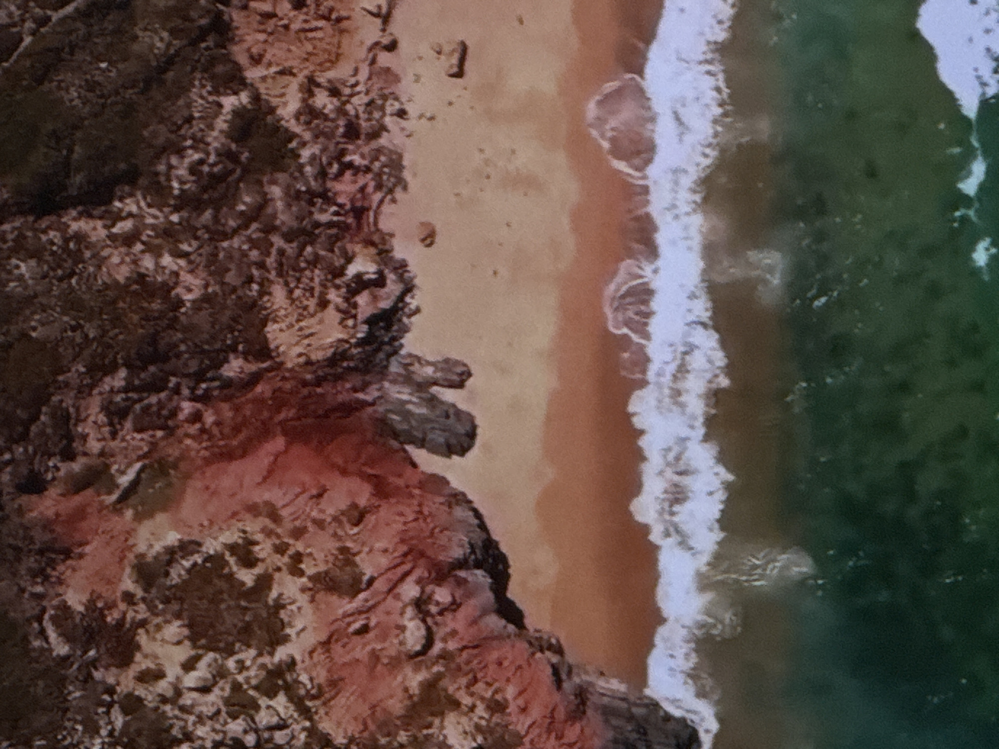 This is an aerial, top-down view of a rugged, reddish-brown rocky cliff meeting a sandy beach and then the ocean. The cliff face is textured with various shades of red, brown, and gray, suggesting a rocky, perhaps eroded, formation. The sand is a light, uniform tan color, with a few scattered dark specks that could be pebbles or debris. The transition from sand to water is marked by a line of white-capped waves breaking on the shore. The ocean water itself is a deep, dark green, with lighter green patches and white foam visible where the waves are forming and receding. The image captures a natural coastal scene without any signs of human activity or urban elements. Therefore, it is not possible to provide details about the location in Sinyavino, Russia, as the image depicts a natural landscape that does not appear to be within a city.