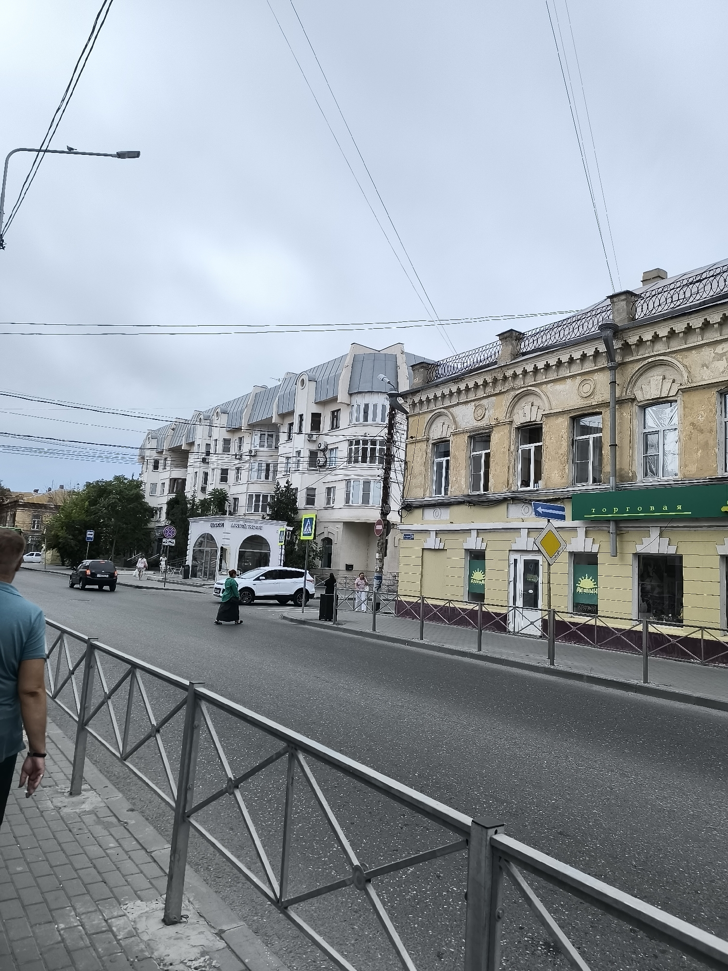 The image shows a street scene in Astrakhan, Russia. In the foreground, a person wearing a teal t-shirt and dark pants is walking on a paved sidewalk with a metal railing. The sidewalk is bordered by the street, which has a gray asphalt surface.
Across the street, a row of buildings lines the road. On the right side, a yellow building with ornate architectural details and arched windows is visible. A green sign with Cyrillic text reads "Торговая" (Torgovaya, meaning "Trading"). Below this, the windows display a sun logo with Cyrillic text.
Further down the street, a modern white building with a mansard roof and numerous windows dominates the left side. Several cars are on the street, including a black car and a white SUV. Pedestrians are also present, with one person in a green outfit crossing the street. Traffic signs and a pedestrian crossing are visible. The sky is overcast.